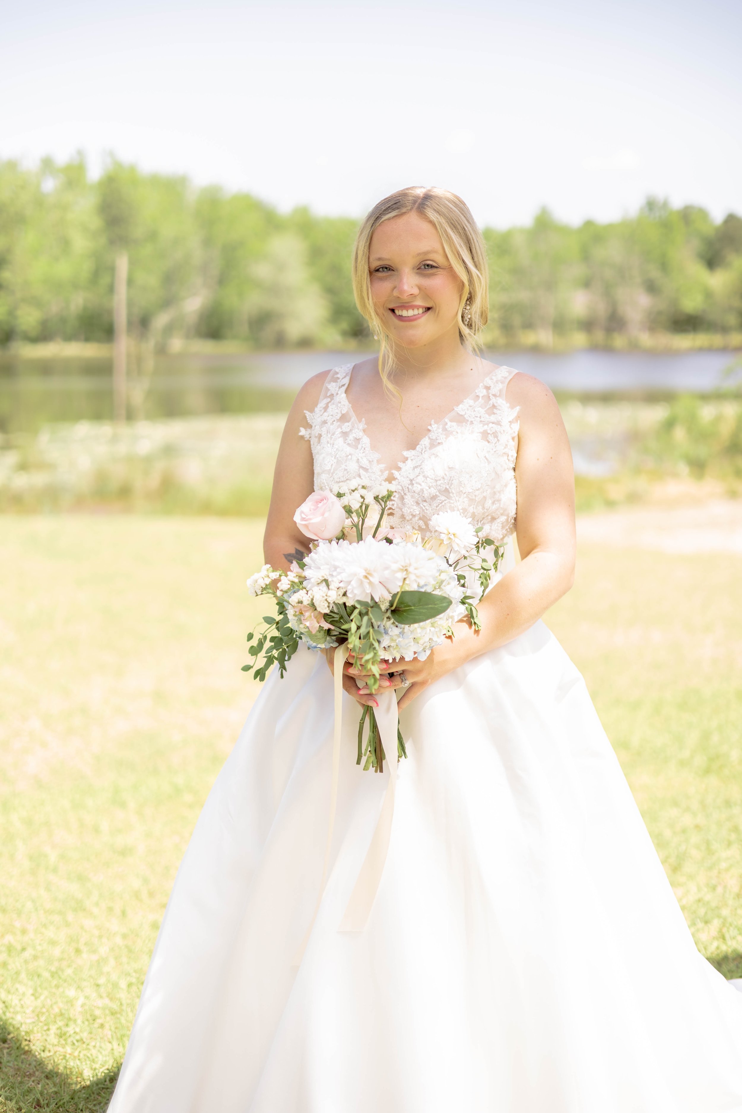 Bride in white gown holding bouquet outdoors with lake and trees in background.
