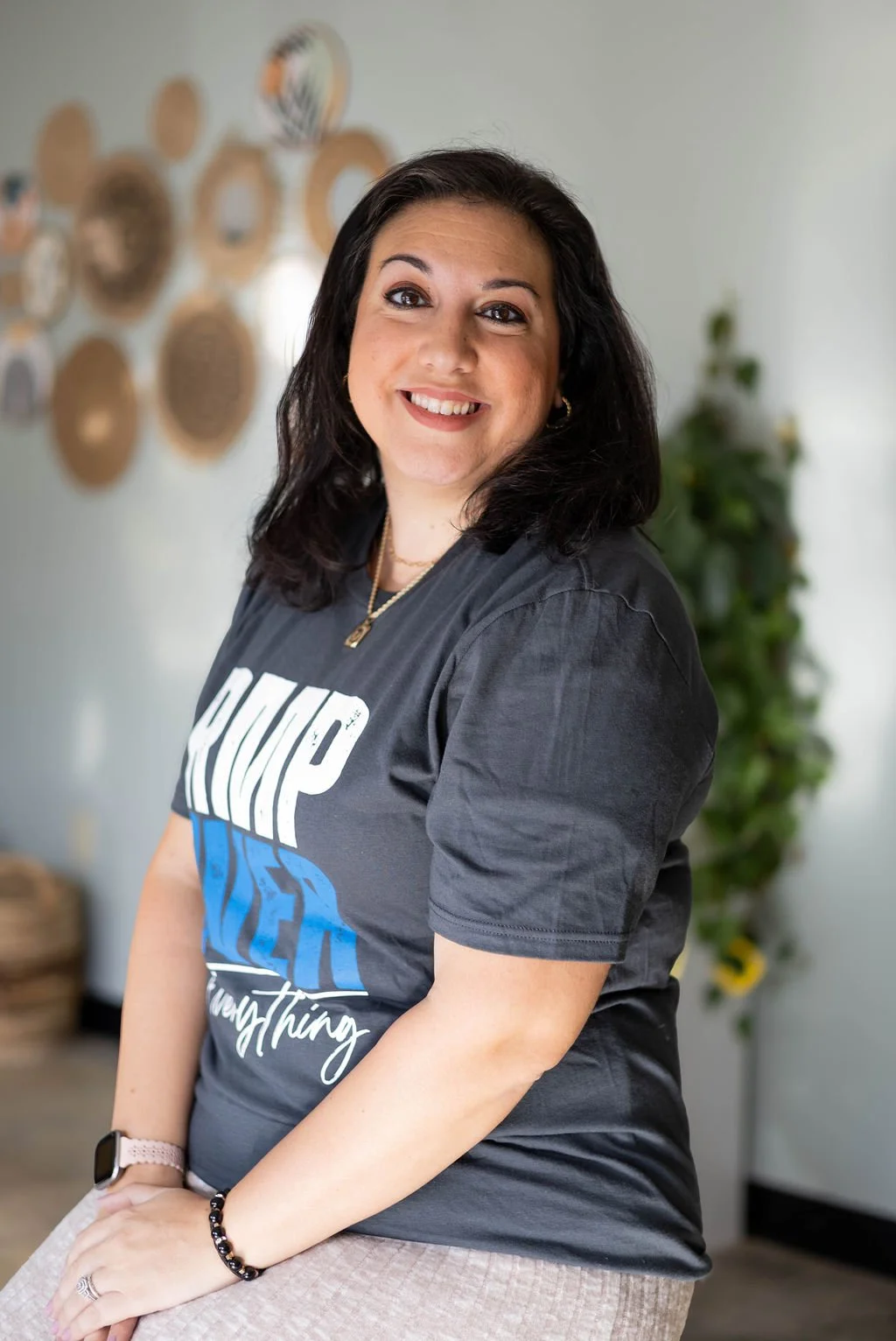 A woman with dark hair and a bright smile, wearing a dark t-shirt, standing in a modern indoor setting with wall decorations and greenery in the background.
