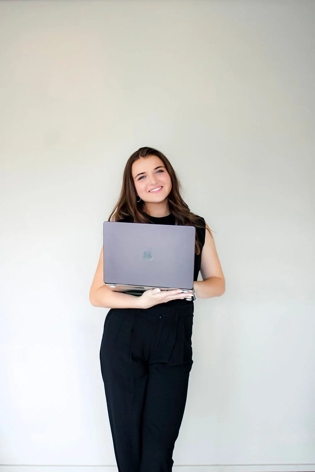 Young woman with brown hair and a black sleeveless top, holding a silver laptop, smiling against a plain light-colored wall.