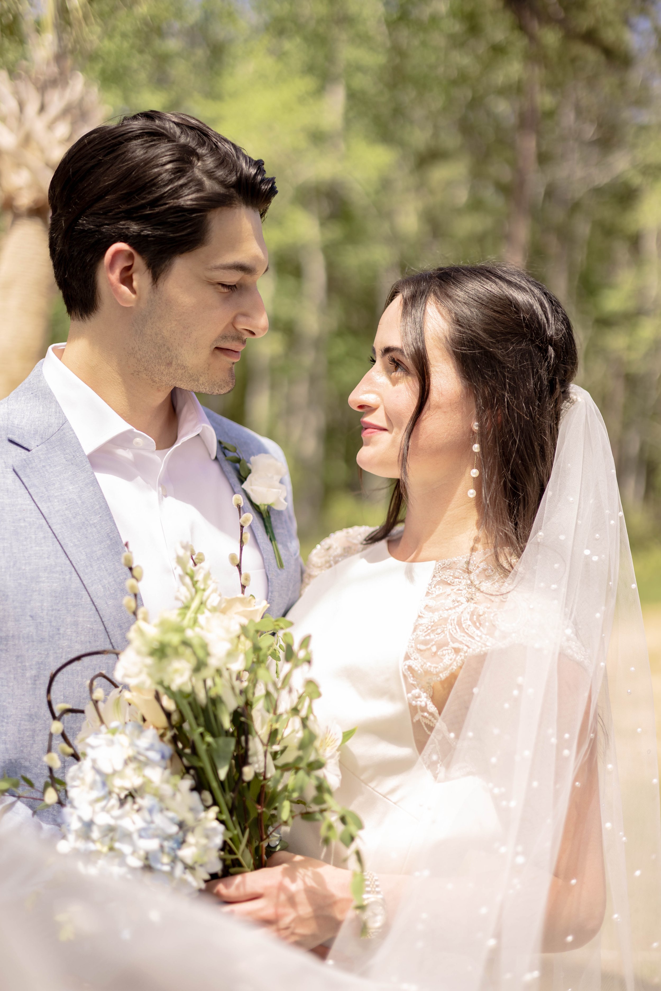 Bride and groom smiling at each other outdoors, the bride holding a bouquet of white and blue flowers with greenery, wearing a veil and lace dress, groom in a light blue suit jacket.