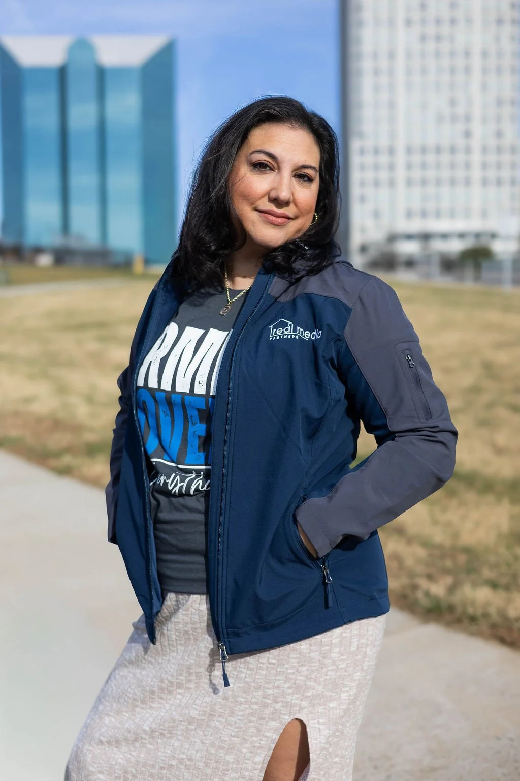 A woman with dark hair, standing outdoors in front of tall buildings in Winston-Salem, NC, wearing a blue jacket, a black T-shirt with a graphic, and a beige skirt, smiling and with her hand in her pocket.