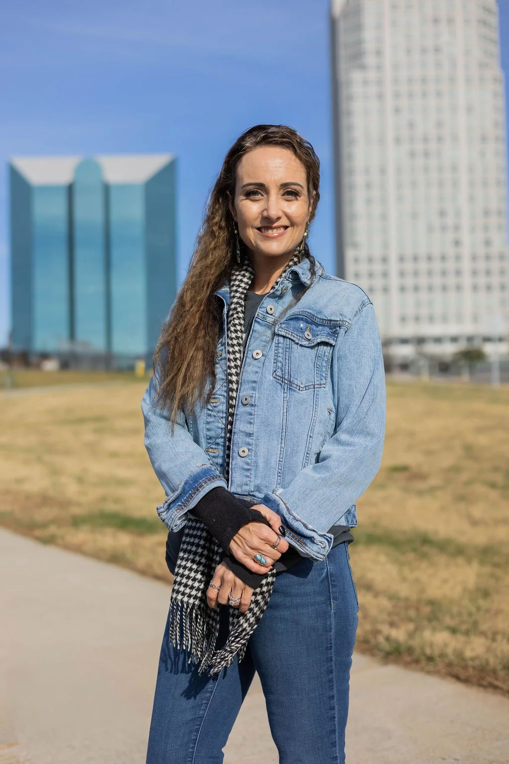 A woman with long curly hair smiling outdoors on a sunny day, wearing a denim jacket and jeans, with tall modern buildings in the background in Winston-Salem, NC.