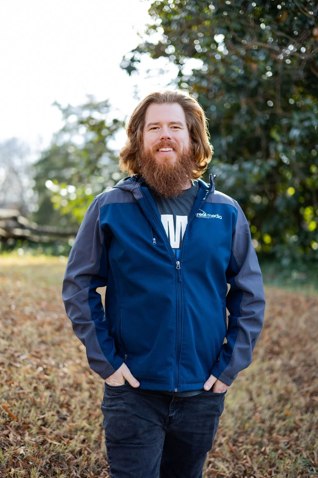 A man with long hair and a beard standing outdoors in a park or backyard, wearing a blue jacket and black pants, with trees and fallen leaves in the background.
