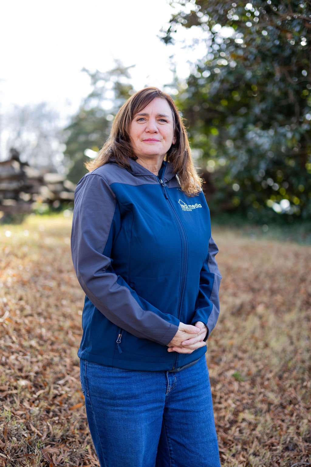 A woman standing outdoors in a park during daytime, wearing a blue and gray jacket and jeans, with hands clasped in front of her, and trees in the background.