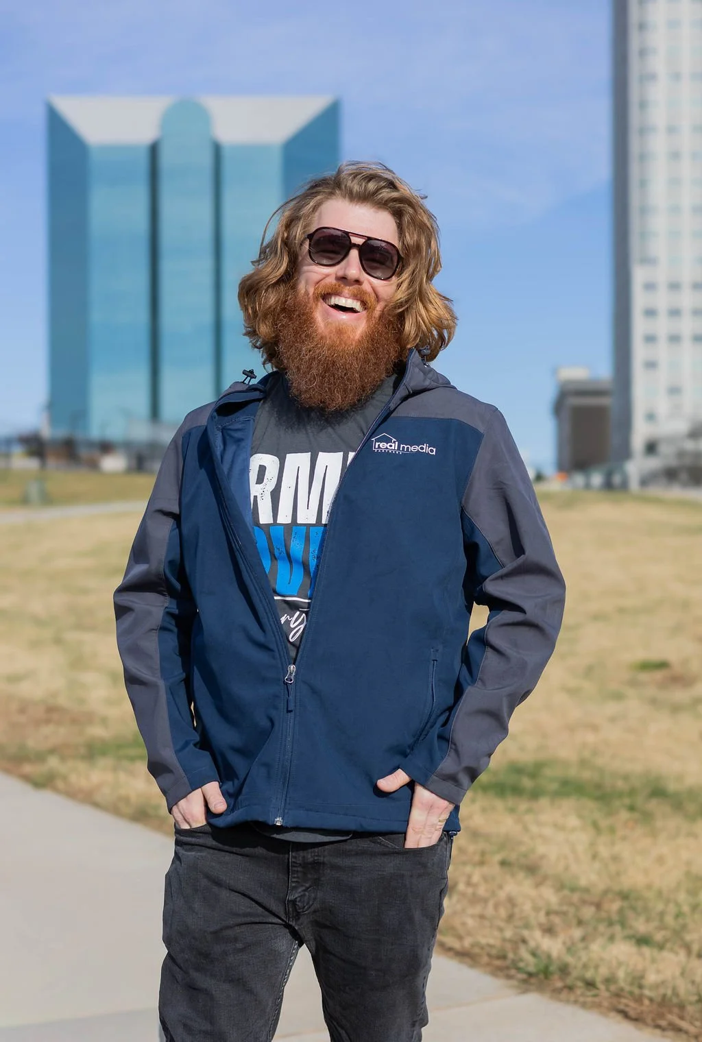 A man with long, wavy hair and a full beard, wearing sunglasses, a navy blue jacket, and a T-shirt, standing outdoors in front of a cityscape with tall buildings in Winston-Salem, North Carolina and a clear blue sky, smiling.