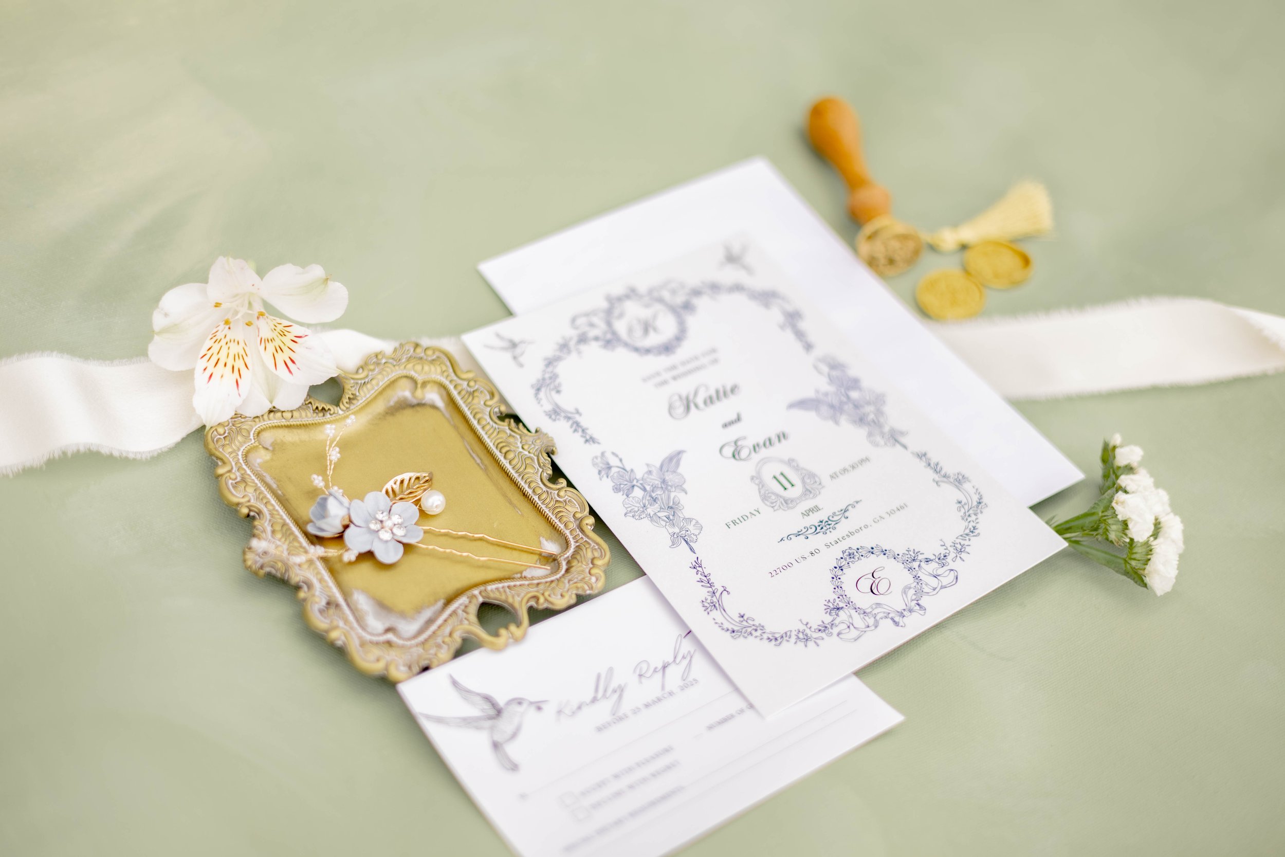 Wedding invitation set with floral design, gold tray with decorative hairpins, white flower, and sealing wax stamps on a light green background.
