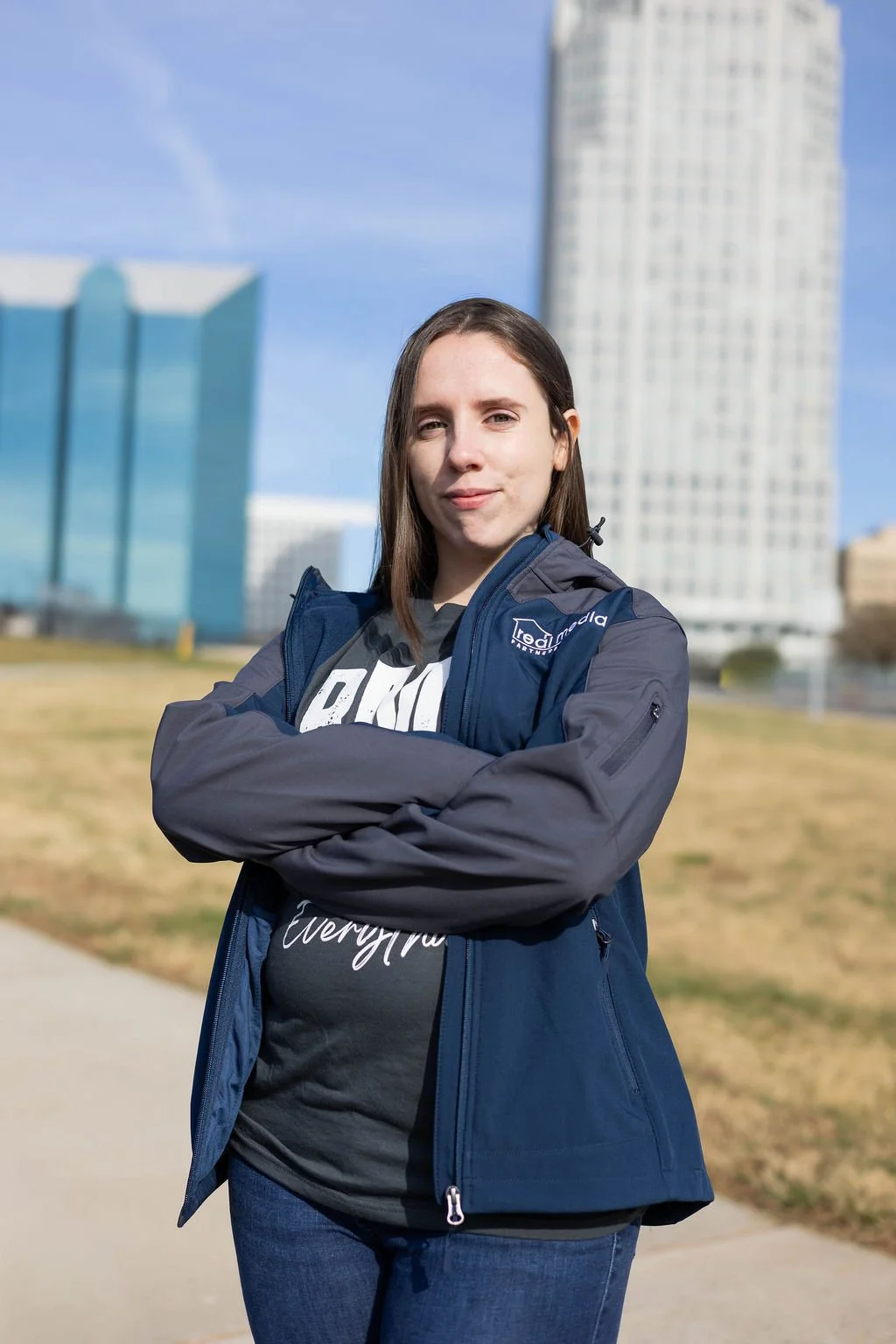 A young woman with brown hair standing outdoors with buildings in the background Winston-Salem, North Carolina, crossing her arms and smiling slightly. She is wearing a black T-shirt and a blue jacket.