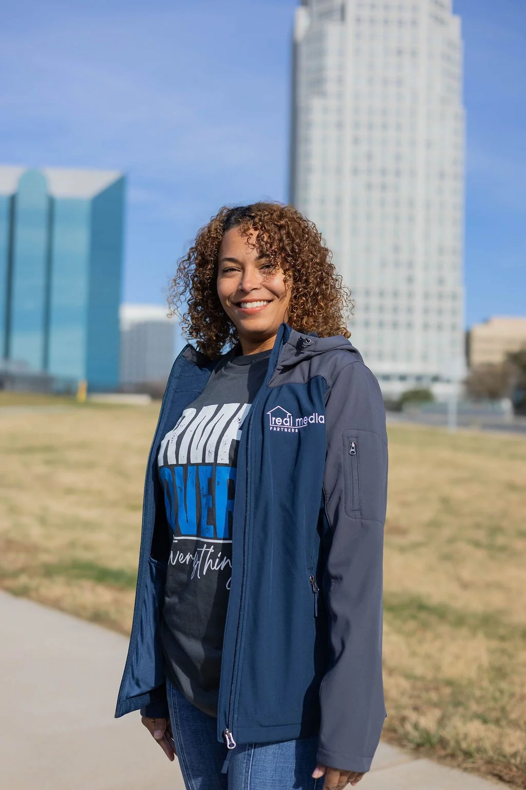 A smiling woman with curly hair standing outdoors in front of city buildings in Winston-Salem, NC, wearing a blue jacket and a black graphic t-shirt.