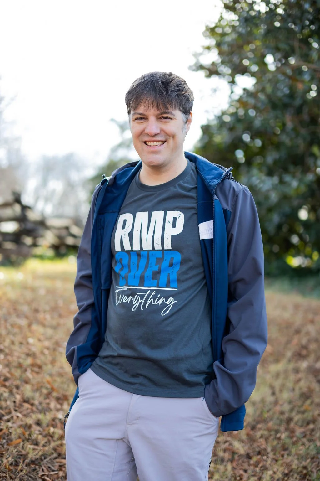 A smiling man outdoors in casual clothing, wearing a gray T-shirt and a blue jacket, standing on a grassy area with trees in the background.
