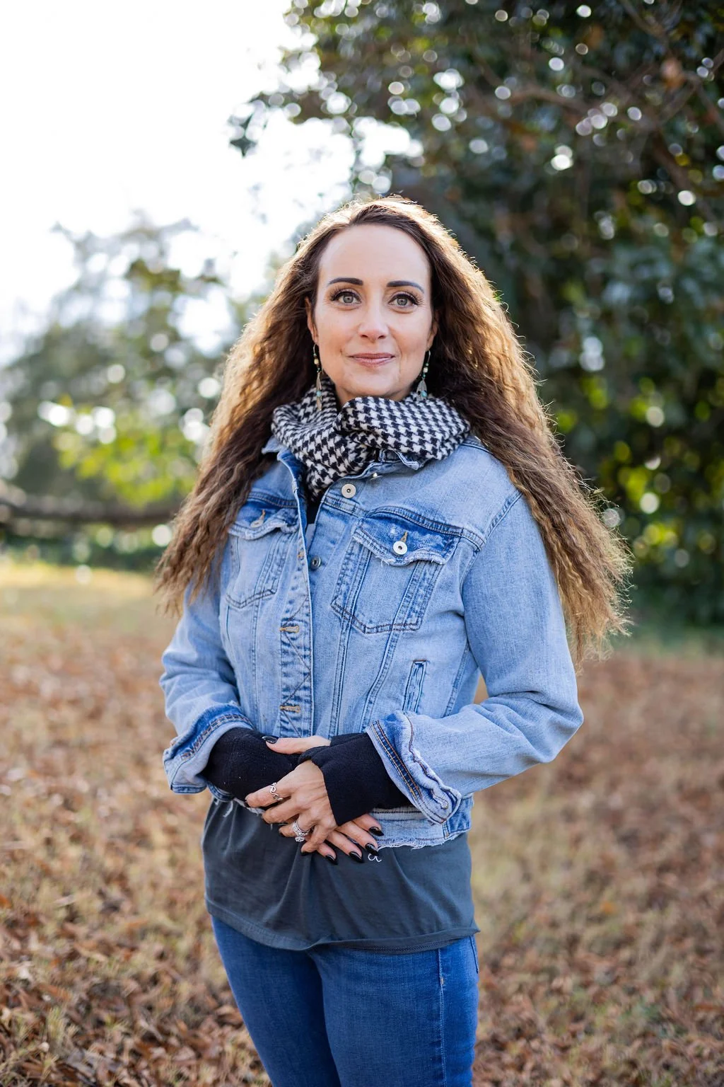 A woman with long, curly brown hair standing outdoors in a park during daytime. She is wearing a denim jacket, a black top layered underneath, a black and white checkered scarf, and black gloves. She has earrings and rings, and is posing with a gentl