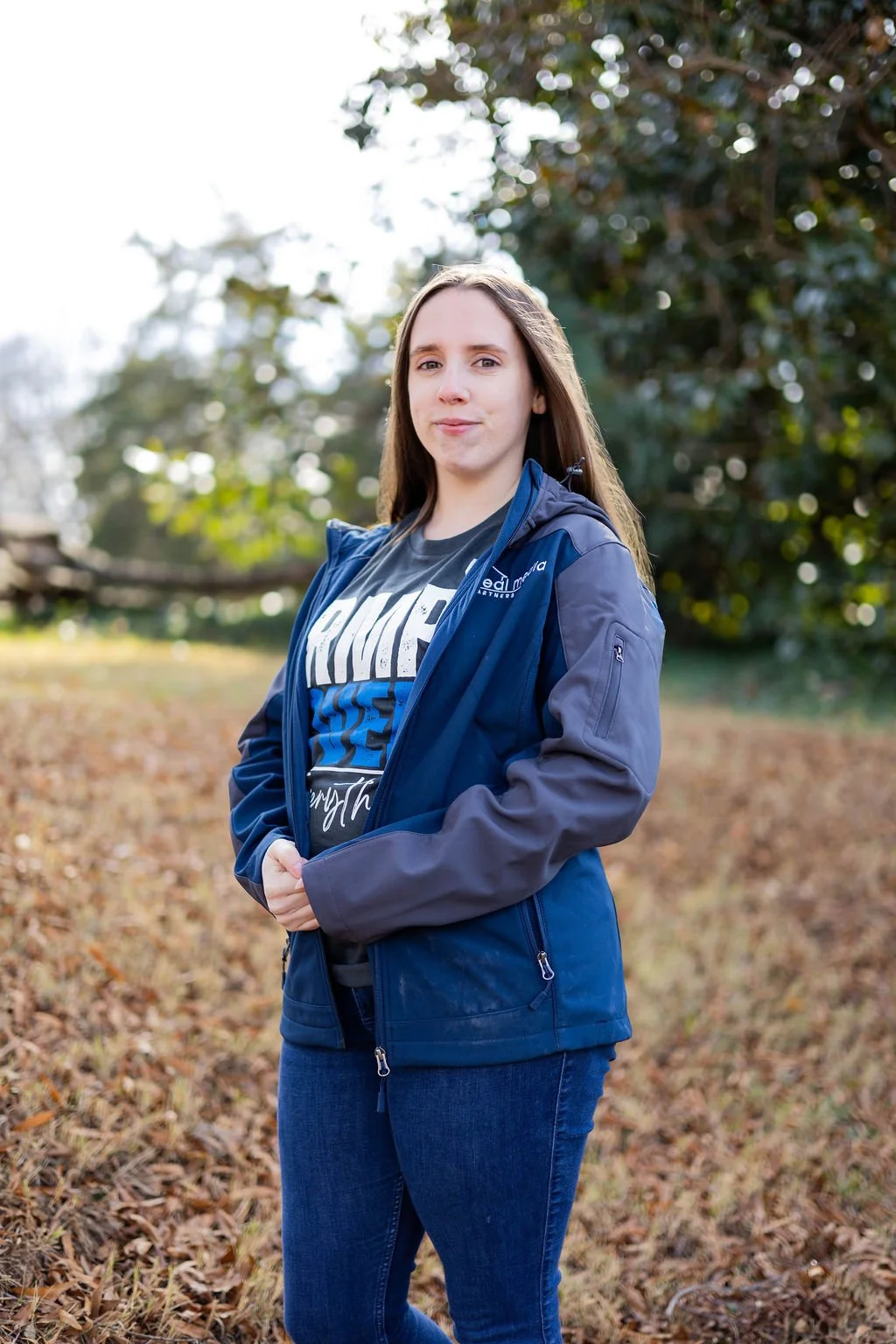 Young woman standing outdoors on a fall day in front of trees, wearing a blue jacket over a black T-shirt with white and blue text, and blue jeans.