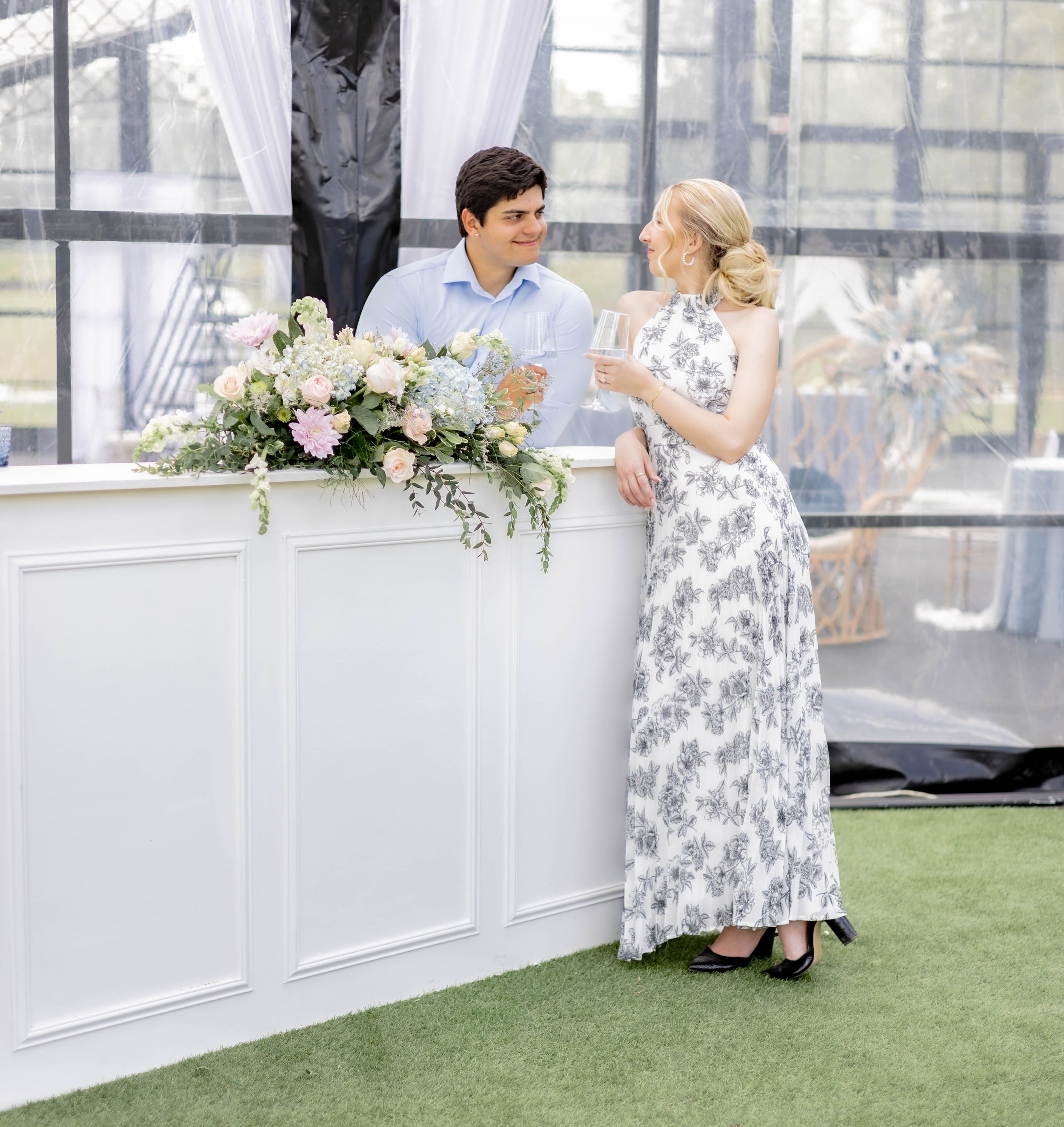 A couple at an elegant outdoor event bar, with a woman in a floral dress holding a drink and a man in a light blue shirt behind the bar. Floral arrangement decorates the counter.
