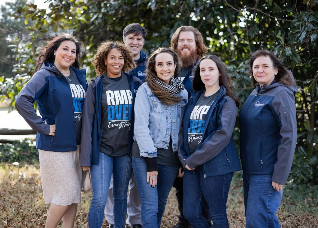 A group of seven diverse people standing outdoors in front of trees, smiling and posing for a photo.