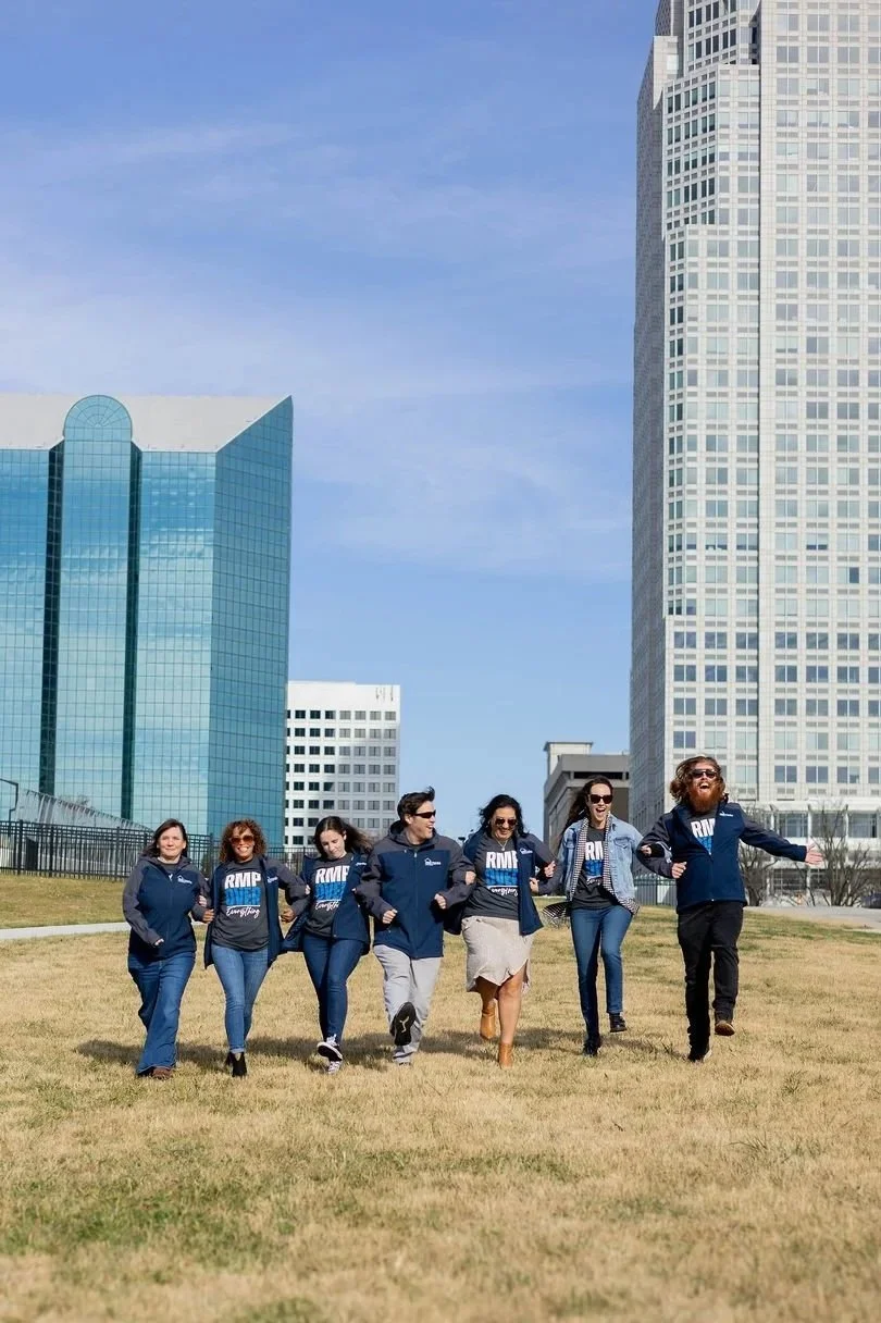 Group of six people walking together outdoors in Winston-Salem, NC with tall office buildings in the background, all wearing casual and team-branded clothing, smiling and enjoying a sunny day.