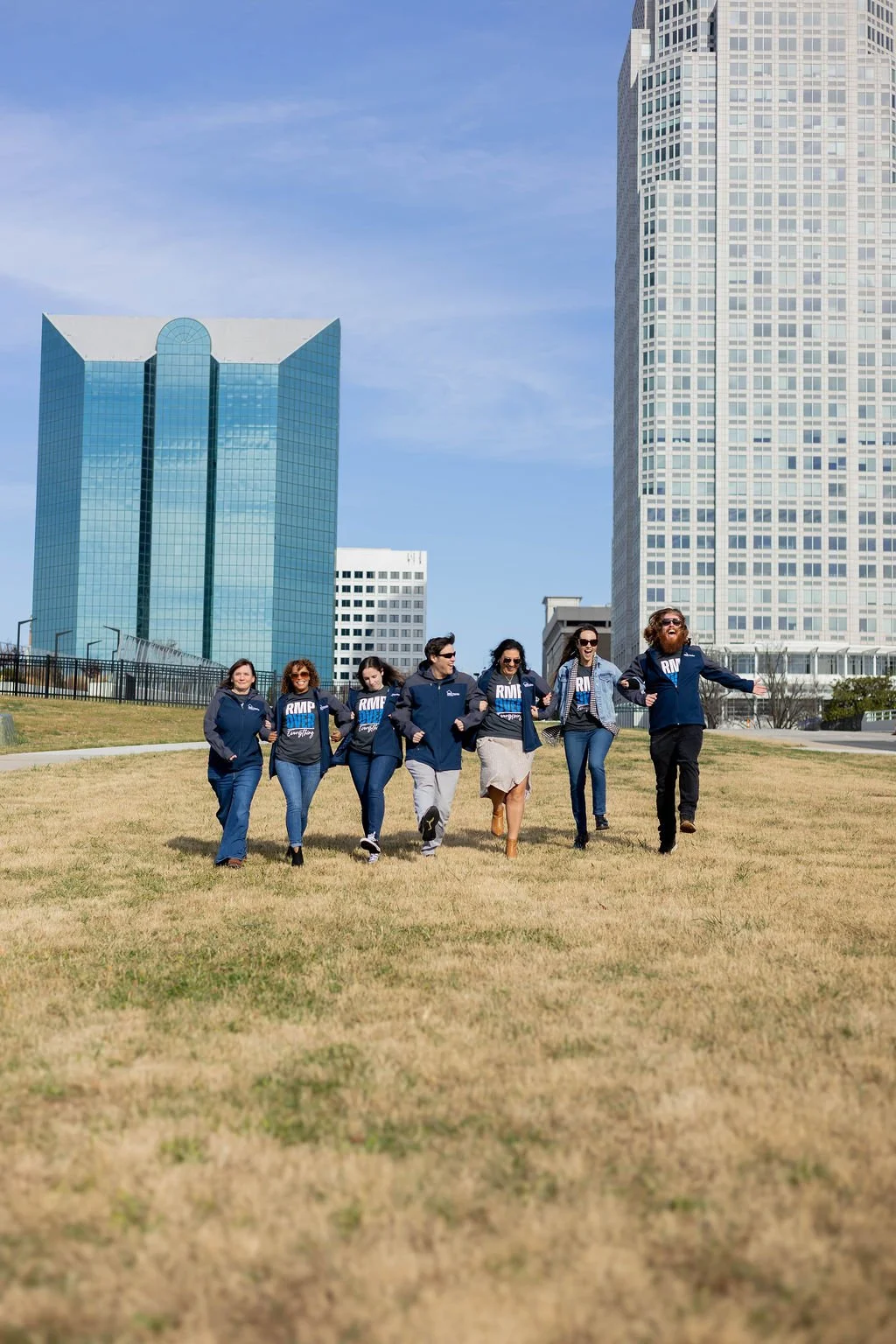 A group of seven people walking together on a grassy area in Winston-Salem, NC with tall modern skyscrapers in the background, wearing matching dark blue jackets and shirts.
