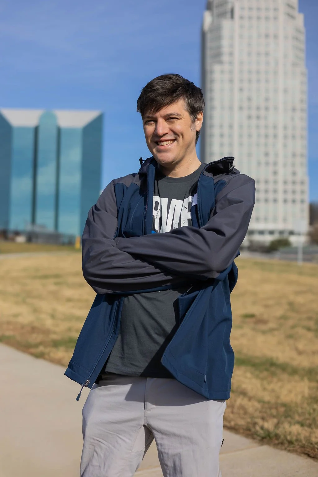 A young man with dark hair wearing a black t-shirt and a blue jacket standing outside on a sunny day with tall buildings in Winston-Salem, NC.