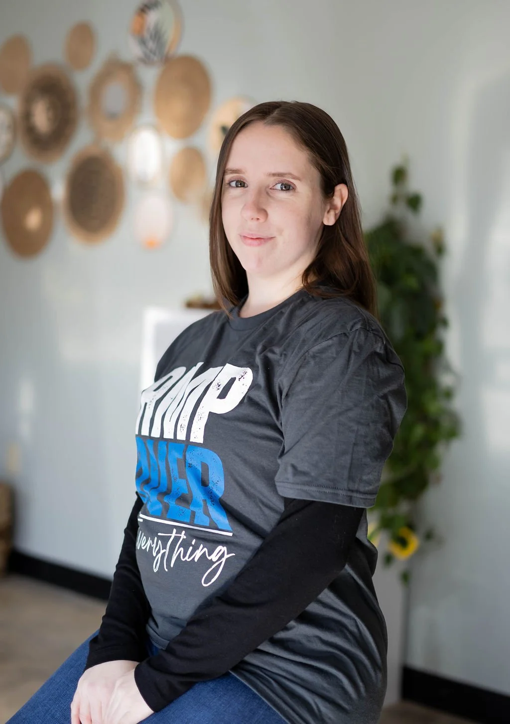 A young woman with brown hair sitting indoors, wearing a dark gray t-shirt with white and blue text, and black sleeves underneath. The background features a decorative wall with various woven circular wall hangings and some green plants.