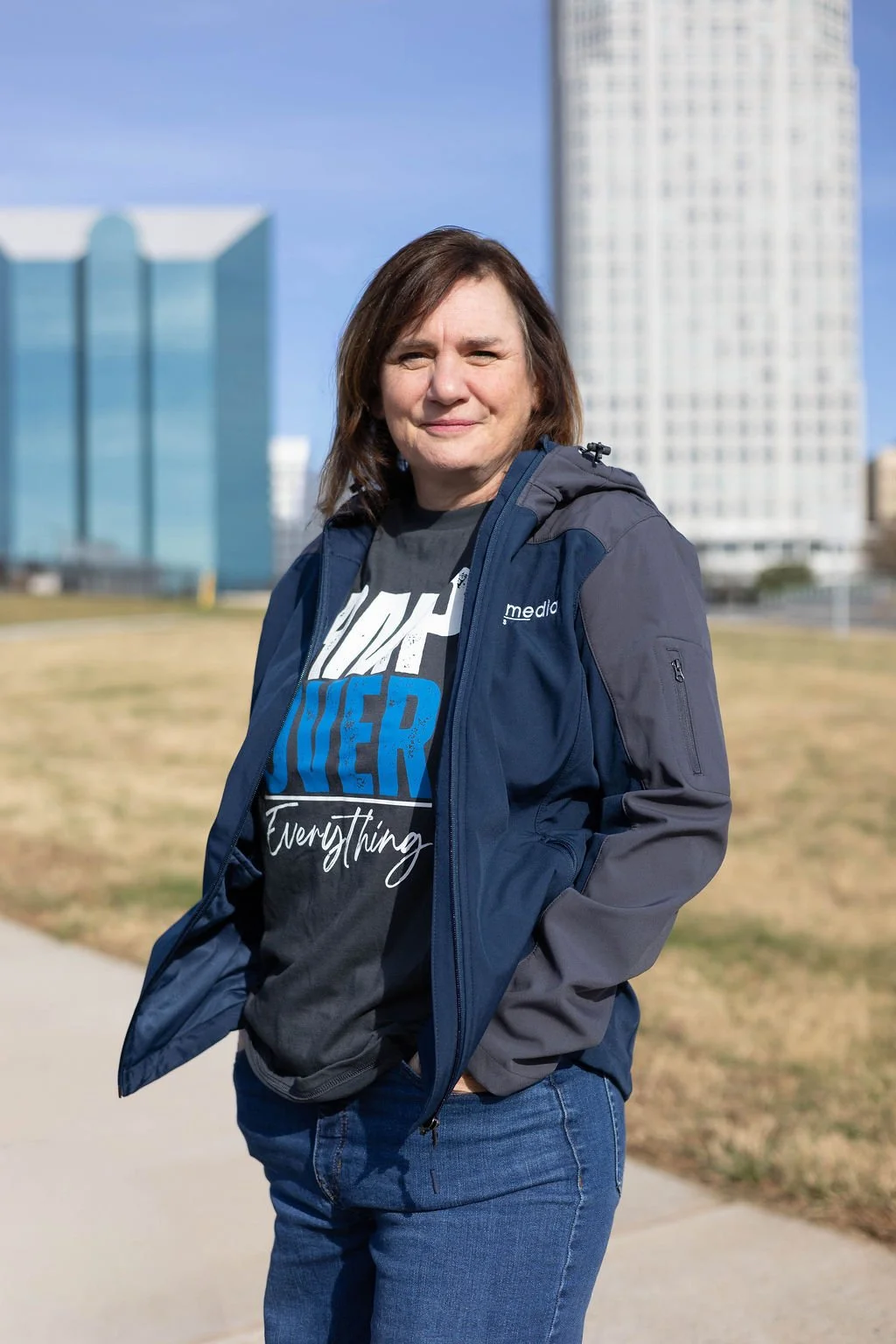 A woman with shoulder-length brown hair and light skin standing outdoors on a sunny day, wearing a dark jacket over a black graphic t-shirt, with a cityscape of tall buildings in the background in Winston-Salem, NC.