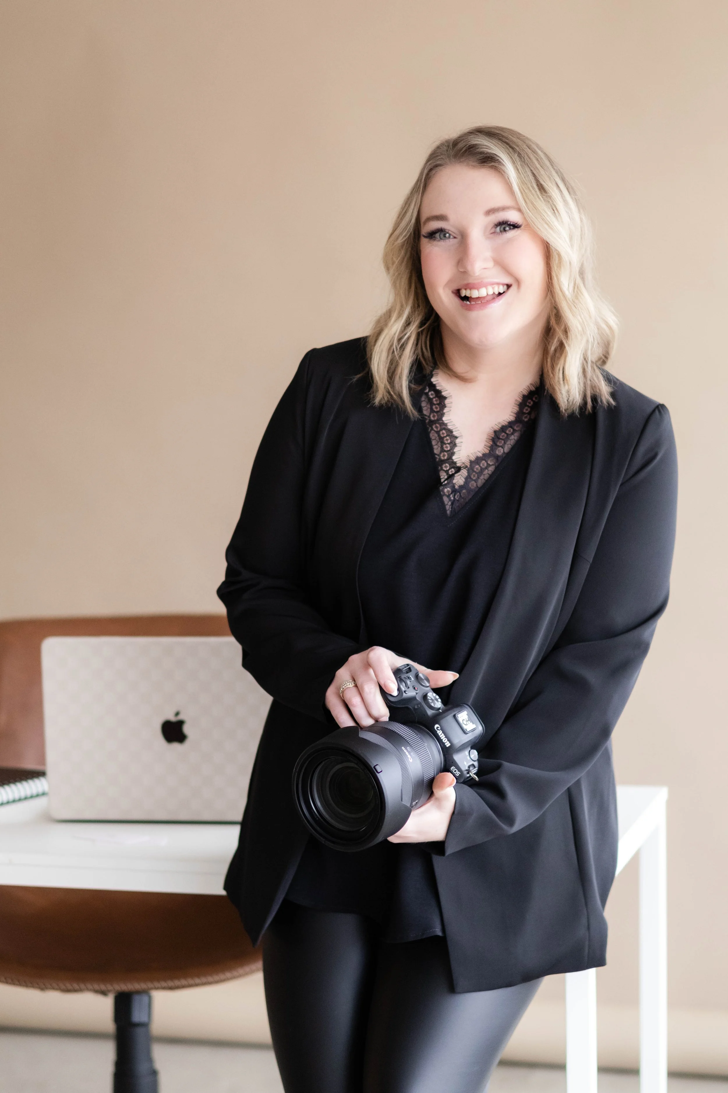 A person smiling, holding a camera, standing next to a table with a laptop.