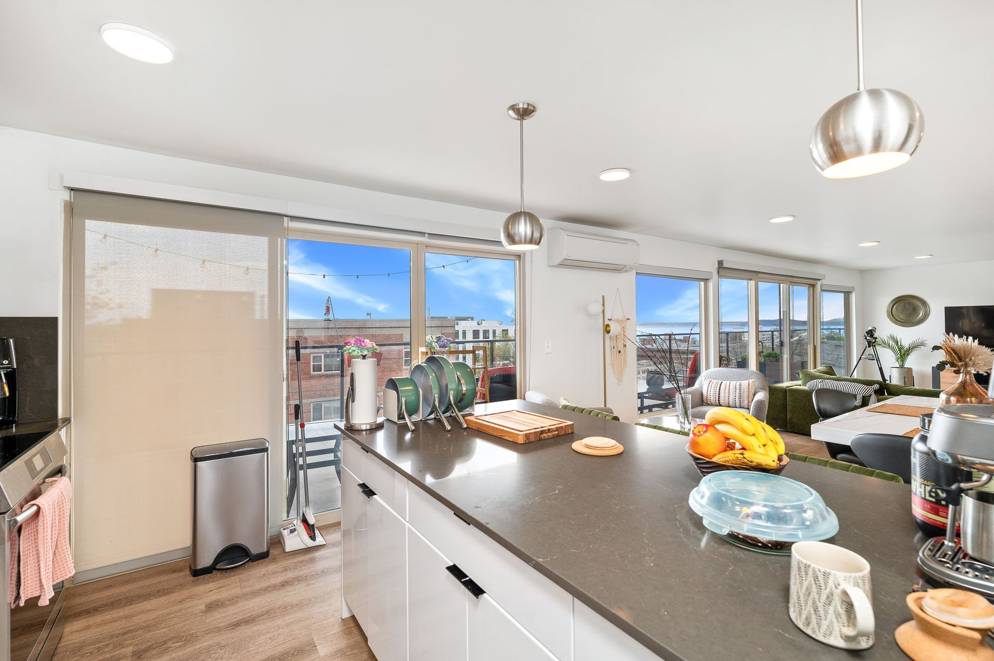 Modern open-plan kitchen and living room with large windows, island counter, kitchen appliances, fruit bowl, and contemporary furnishings. Outdoor view includes patio with seating and neighboring buildings.