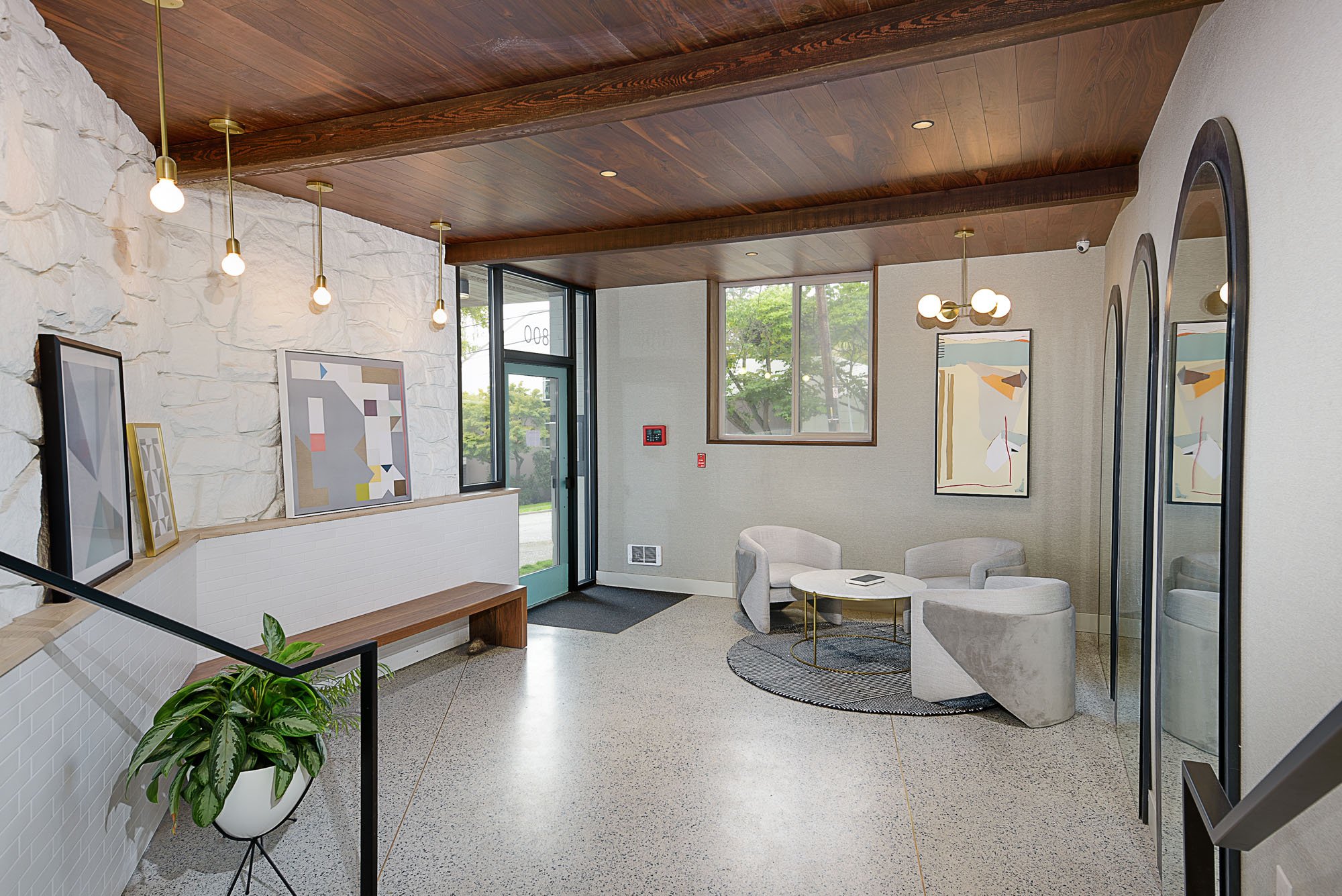 Modern lobby interior with gray armchairs, round table, abstract wall art, pendant lights, wooden ceiling, stone and brick walls, mirrors, and potted plant.