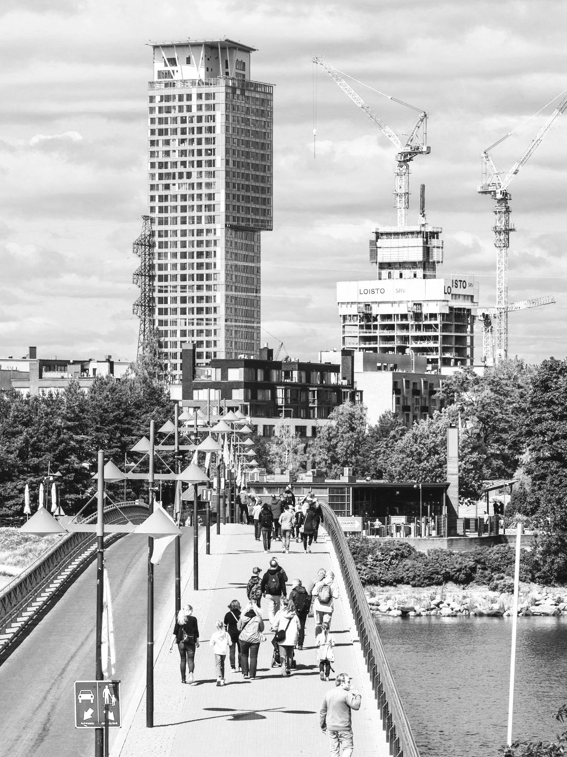 Black and white photo of people walking on a bridge. High-rise buildings and construction cranes in the background.