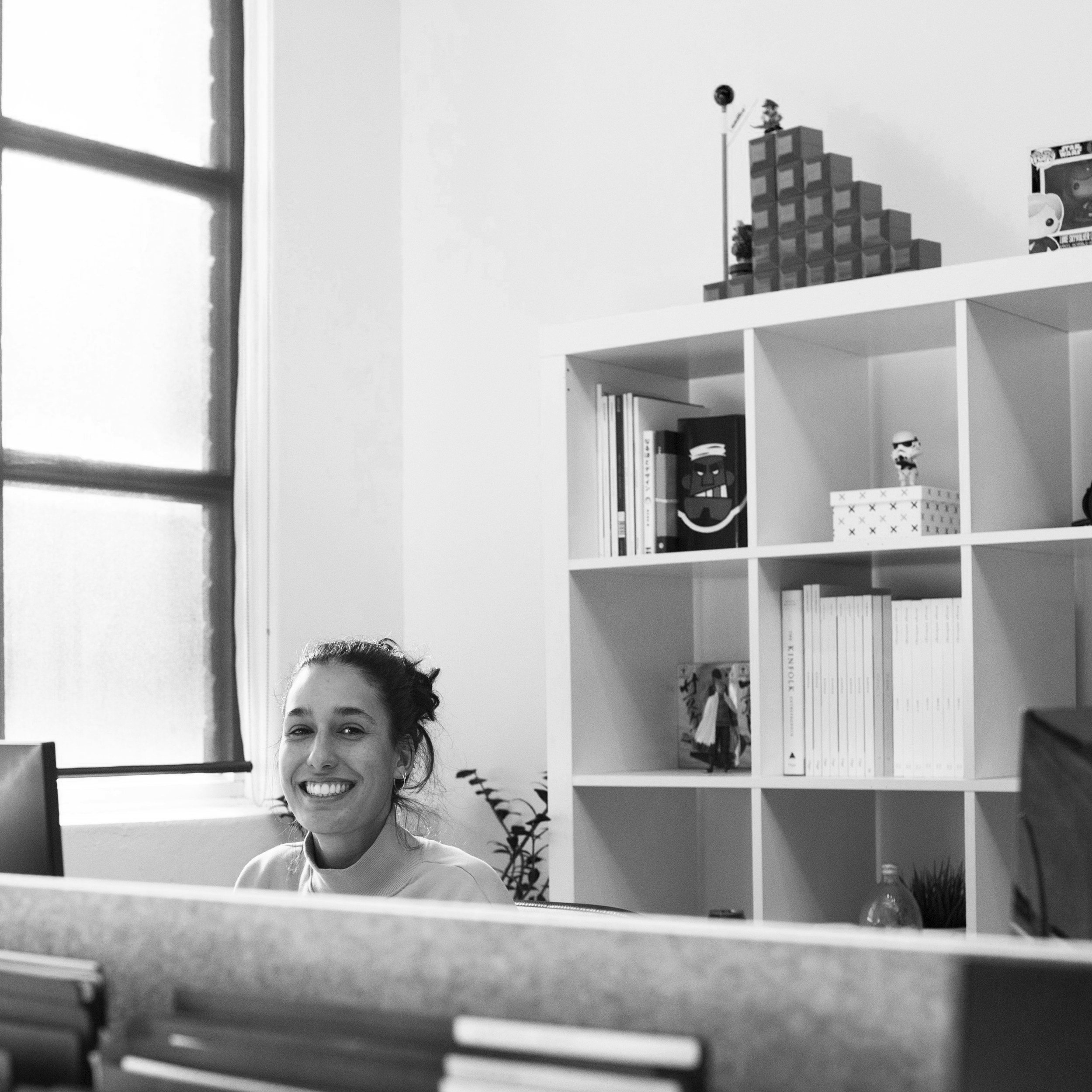 Black and white photo of a smiling woman at a desk in an office, with bookshelves and a window in the background.
