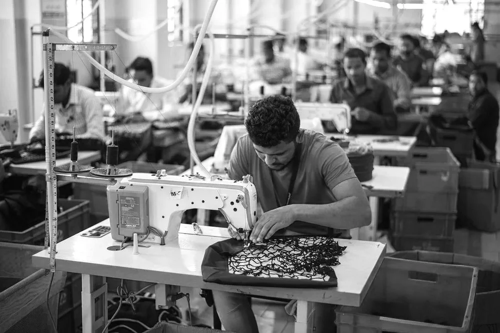 Man operating industrial sewing machine in a busy textile factory with workers in background.
