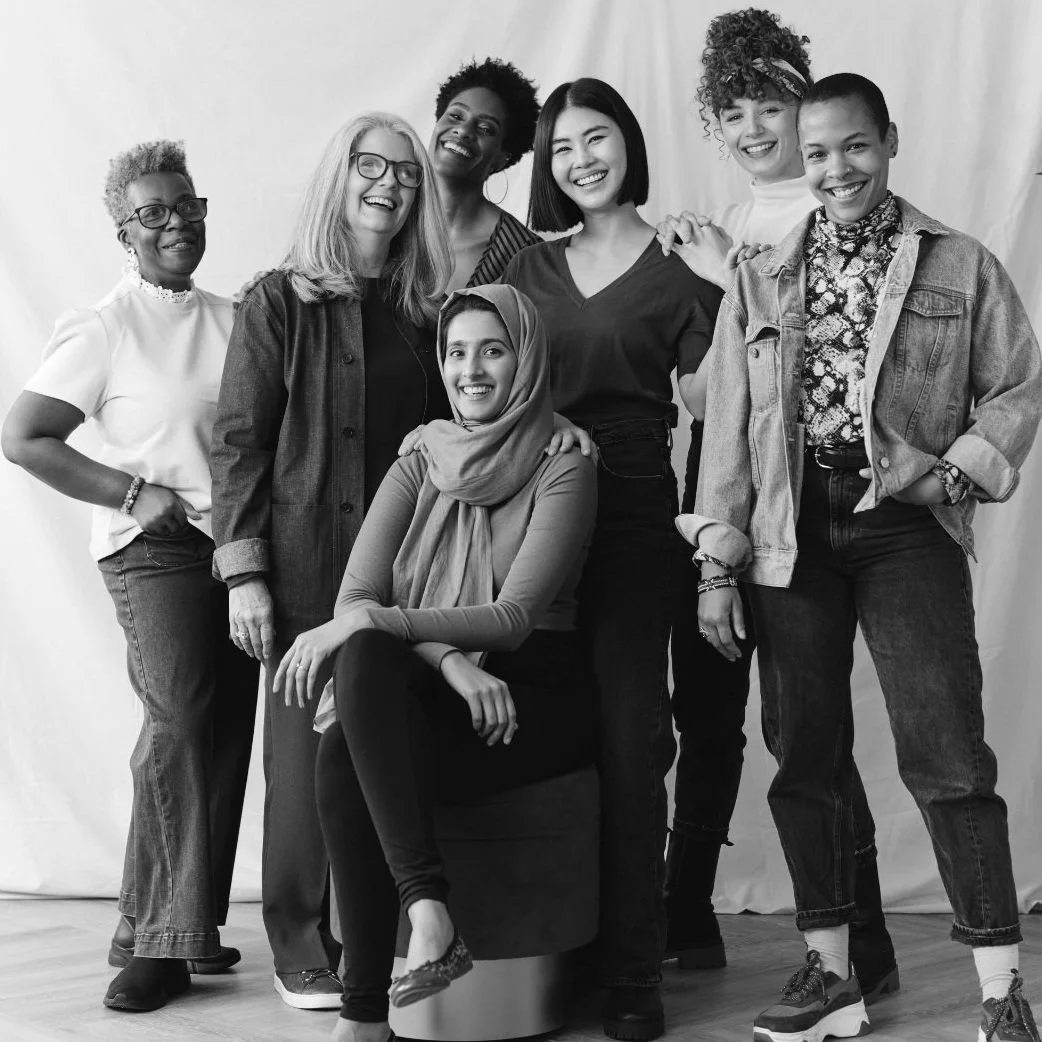 Group of diverse women posing and smiling in a black-and-white photo, with one seated and others standing around her on a light-colored backdrop.