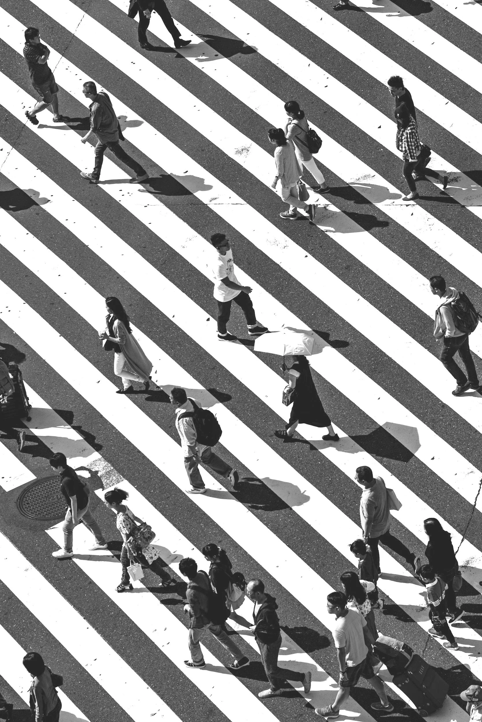 Aerial view of people walking across a wide, striped pedestrian crosswalk in black and white.
