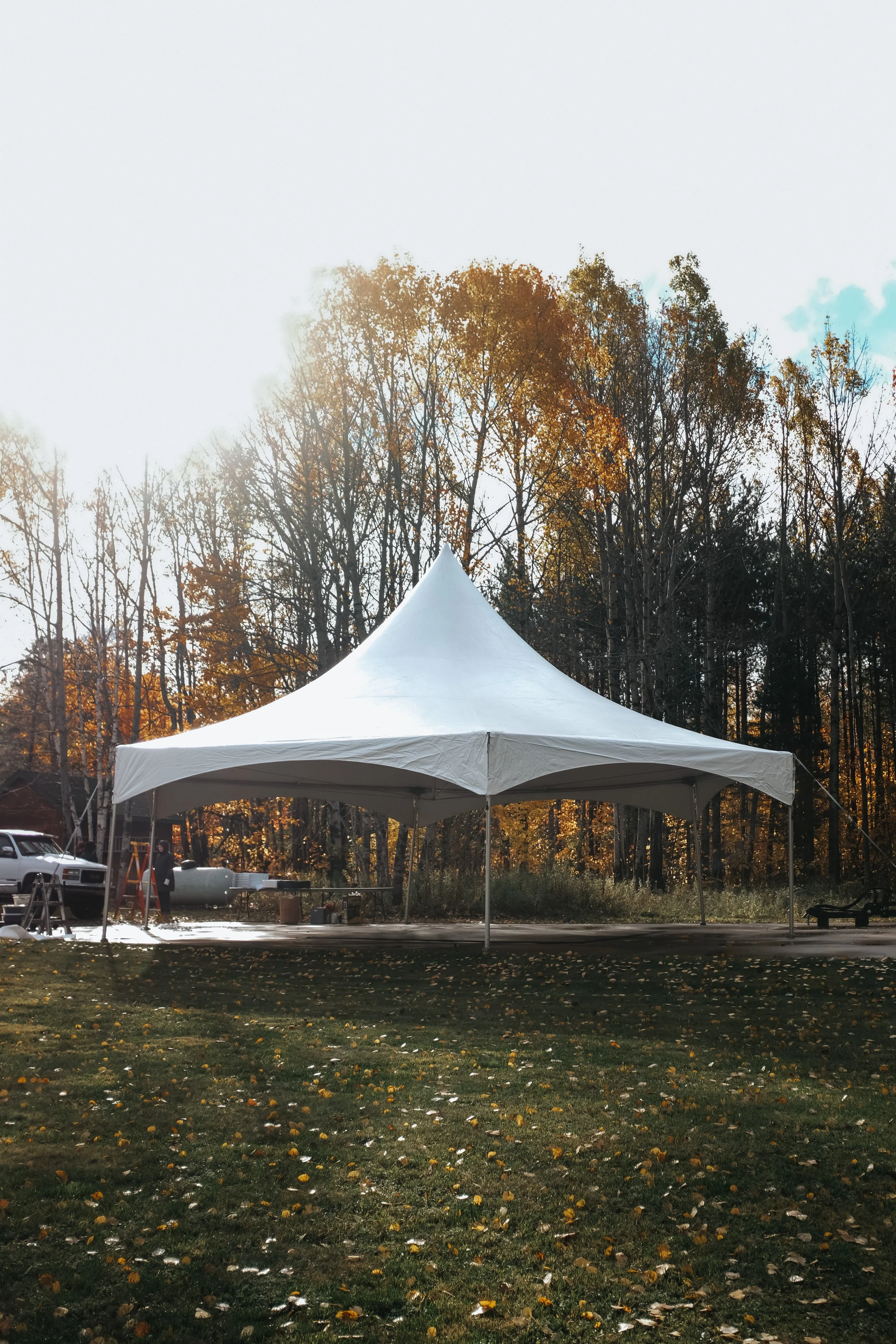 A white outdoor tent set up on grass with autumn trees in the background and vehicles nearby.