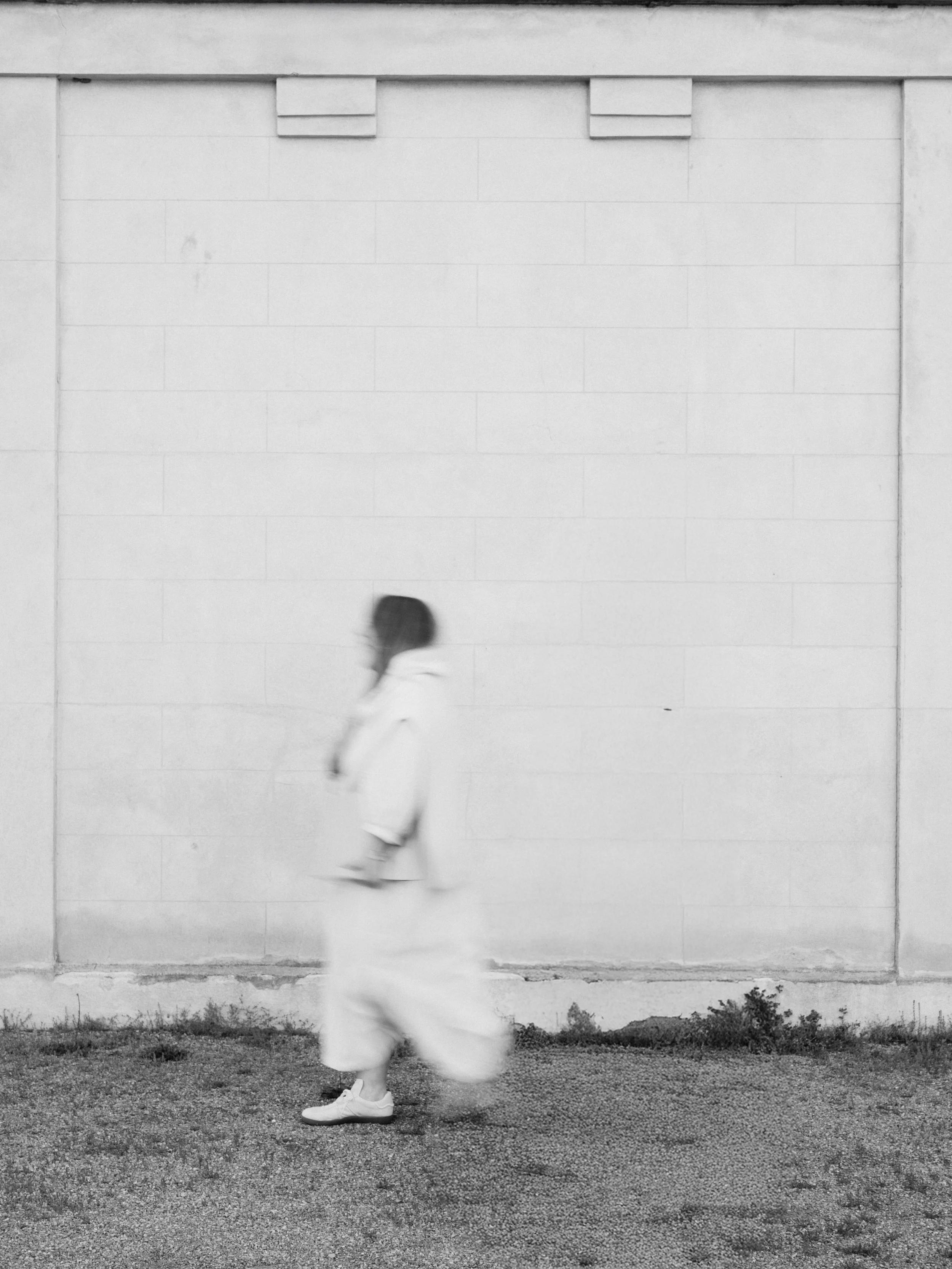 A person walking past a plain white wall in black and white, with the person slightly blurred.