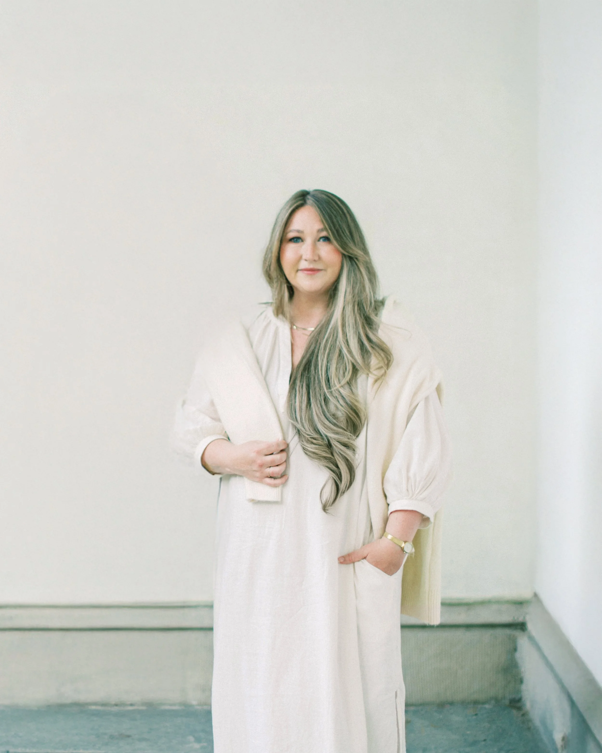 A woman with long, wavy blonde hair wearing a cream dress and a matching cardigan, standing indoors against a plain light-colored wall.