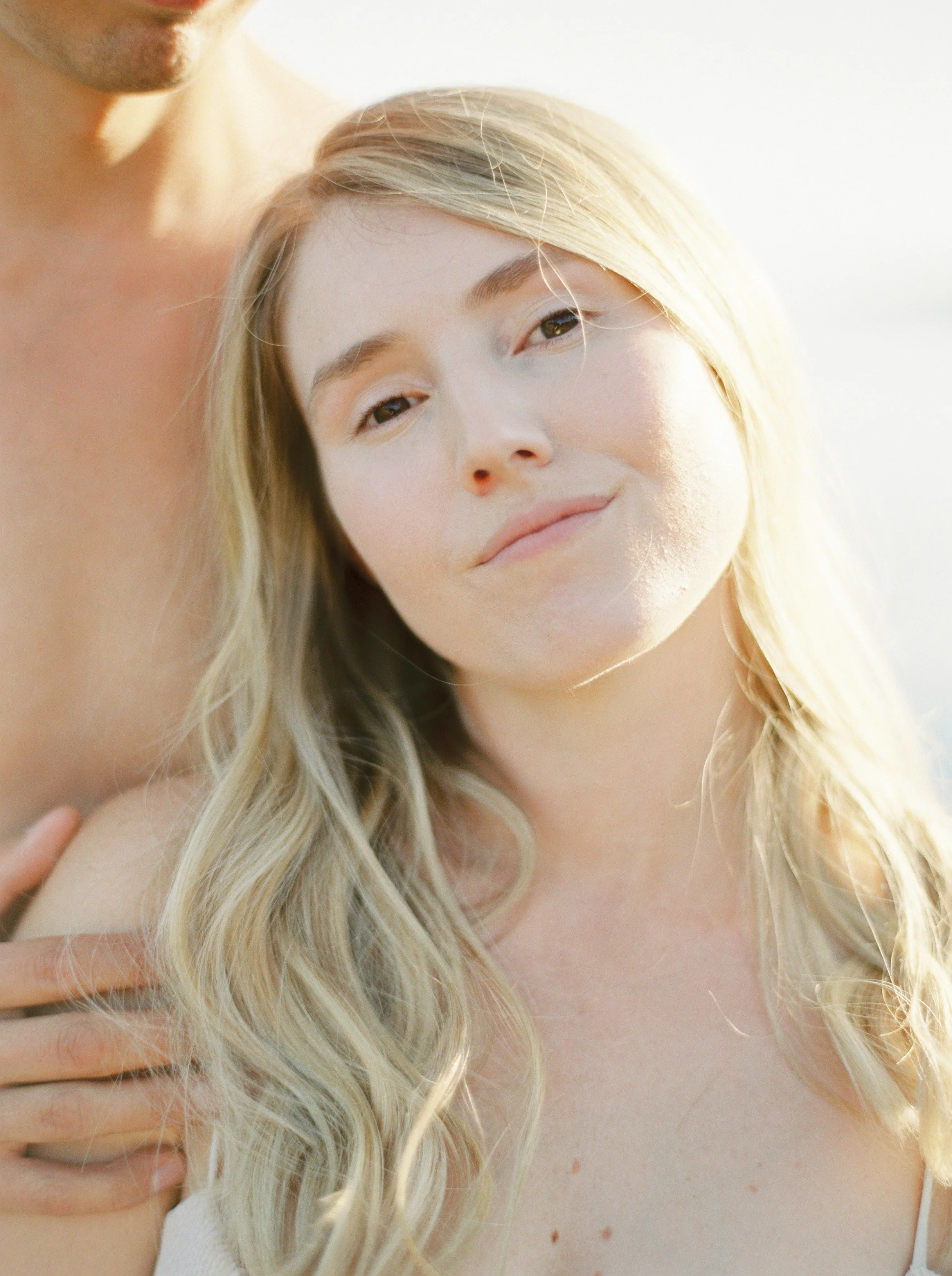 A close-up of a woman with long blonde wavy hair and a man partially visible behind her, outdoors with bright natural lighting.