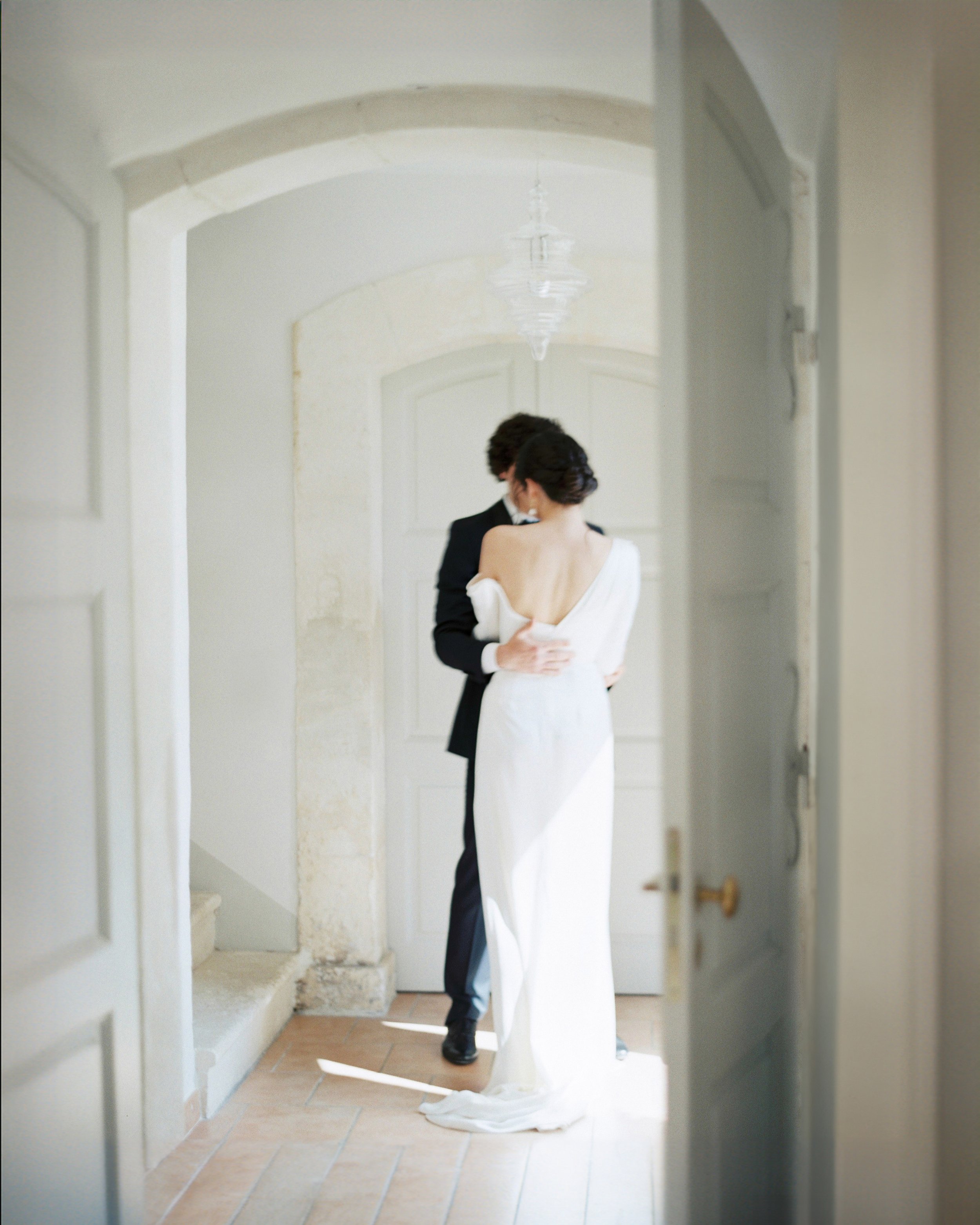 A couple dressed in wedding attire standing close together in a softly lit room, with the woman wearing a white wedding dress and the man in a dark suit, embracing affectionately.