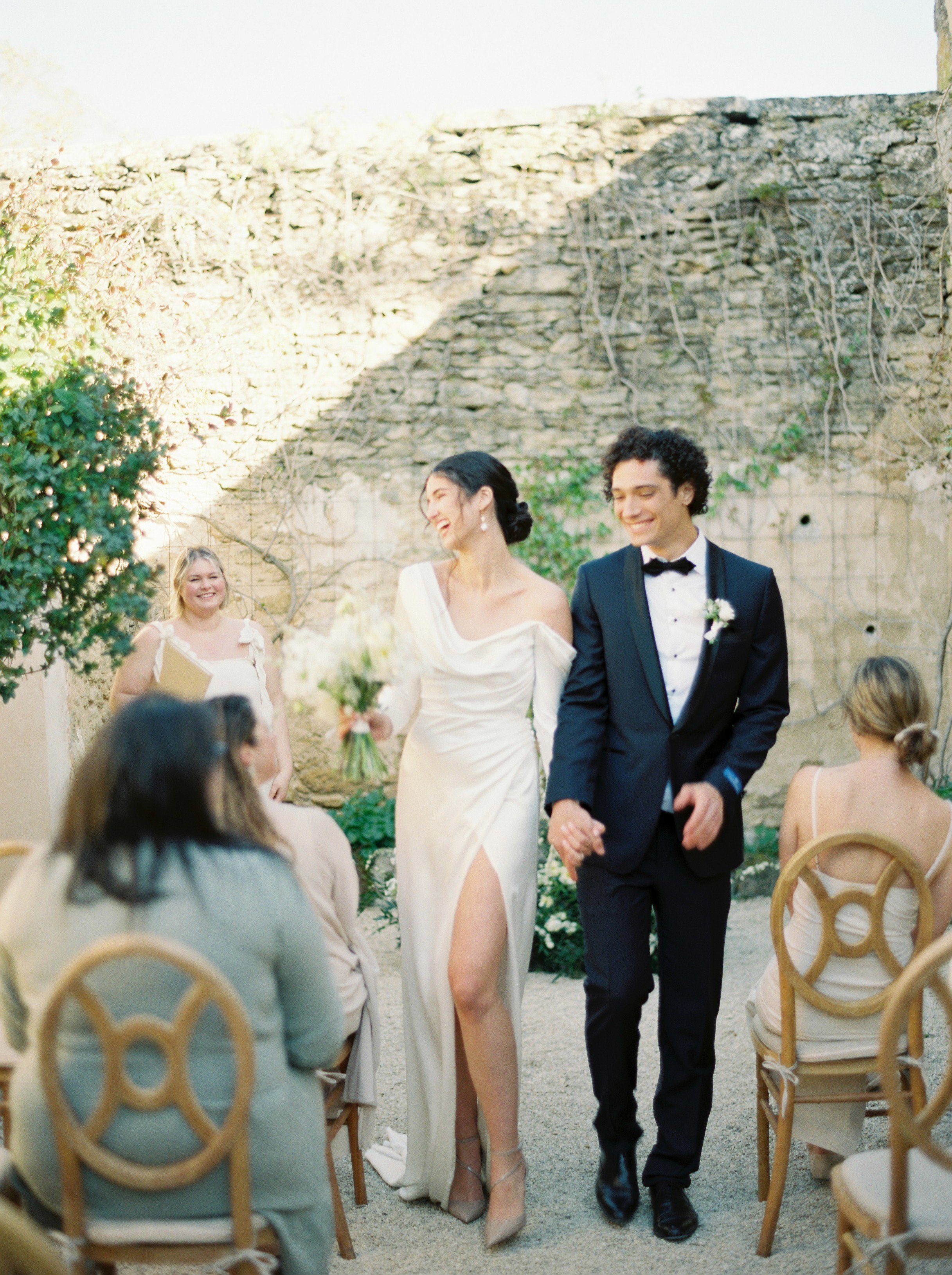 A bride and groom walking hand in hand down the aisle at their wedding ceremony outdoors, with guests seated on either side and a stone wall in the background.
