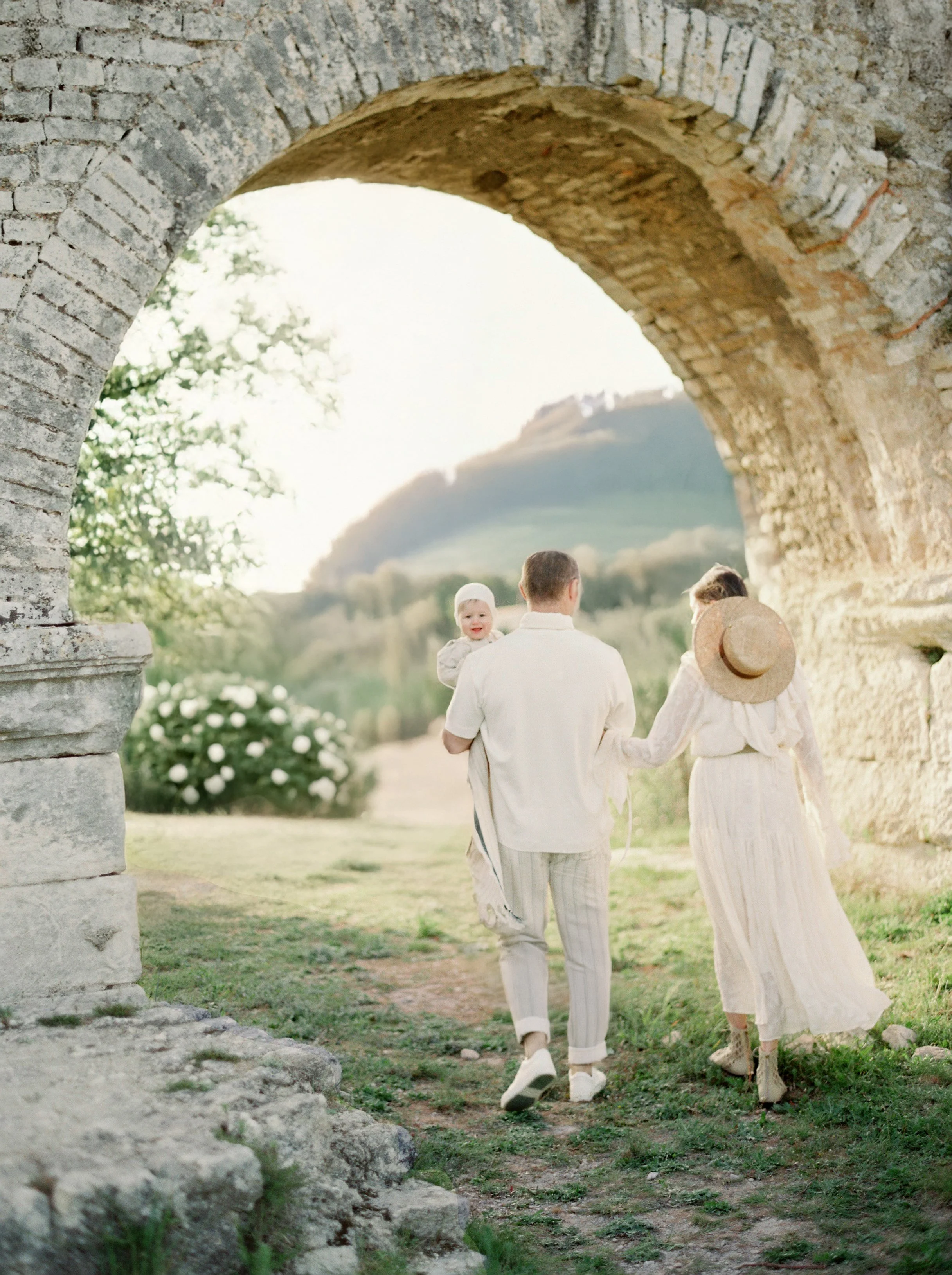 A family walking through an old stone archway in a scenic outdoor setting on a sunny day, with a background of green hills and a clear sky.