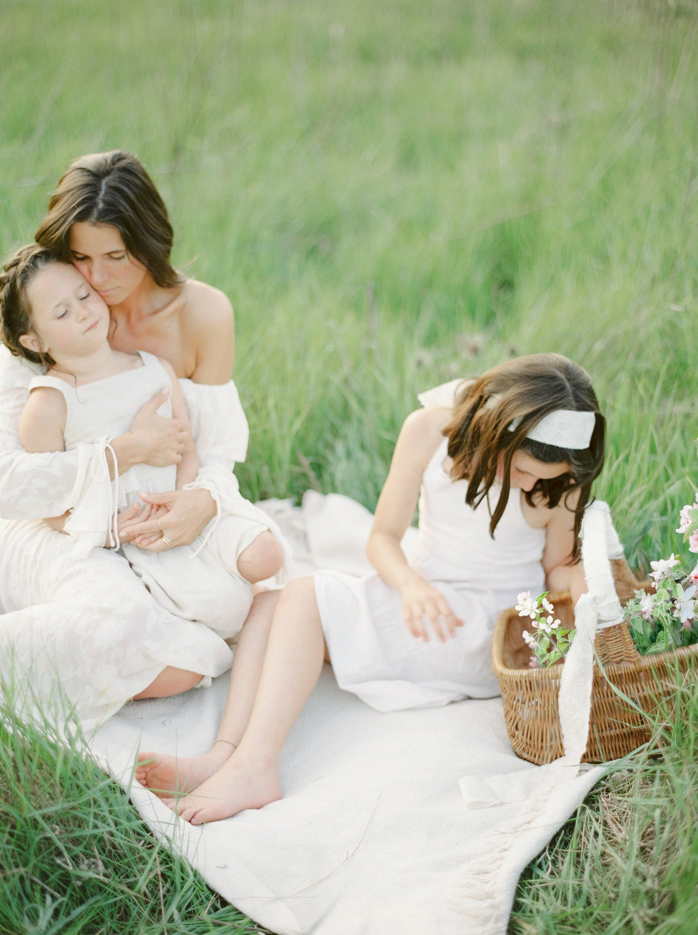 A woman and two young girls having a tender moment outdoors on a grassy field, seated on a white blanket with a basket of flowers nearby.