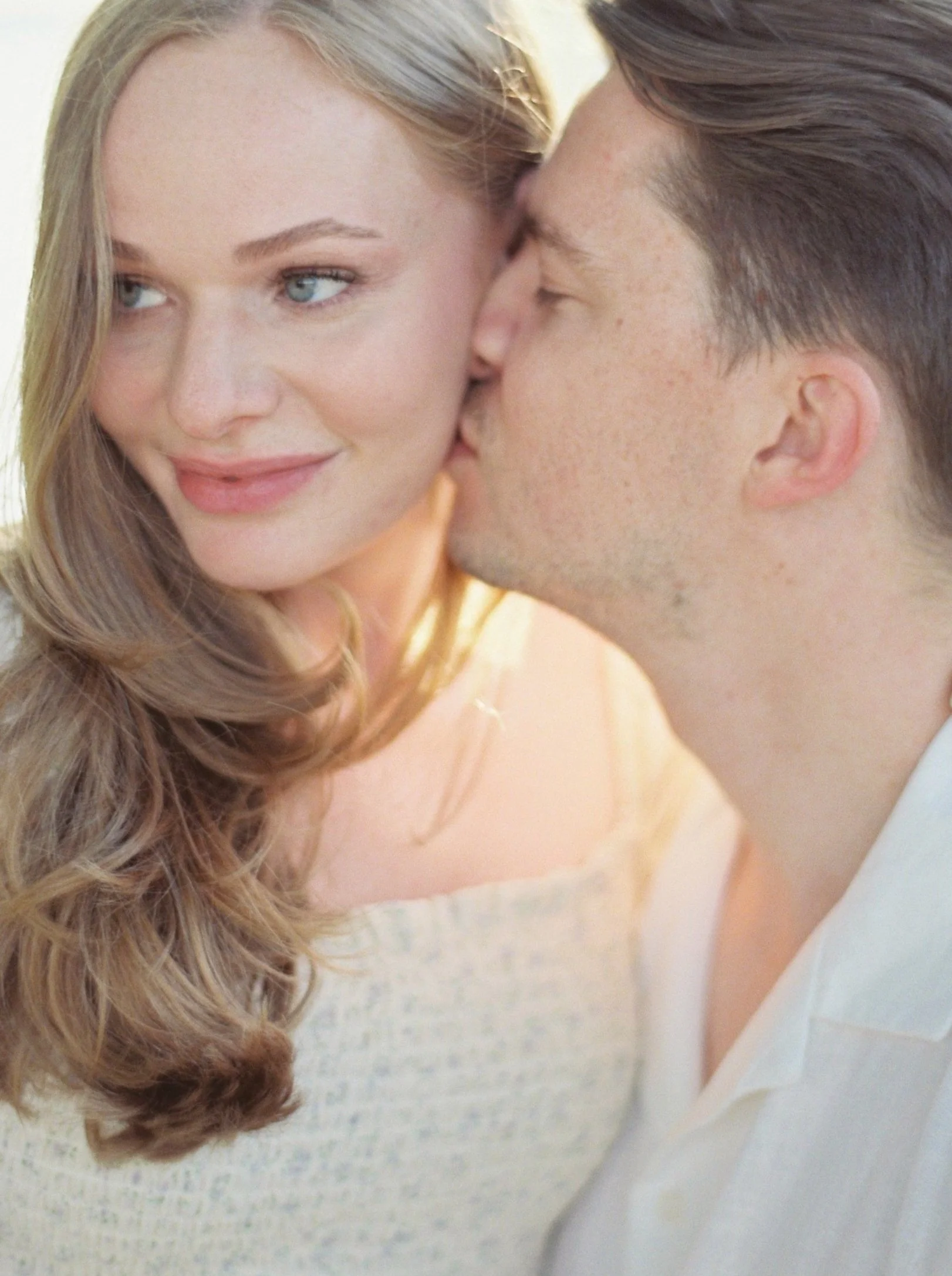 A man is giving a woman a kiss on the cheek, the woman is smiling and looking away.