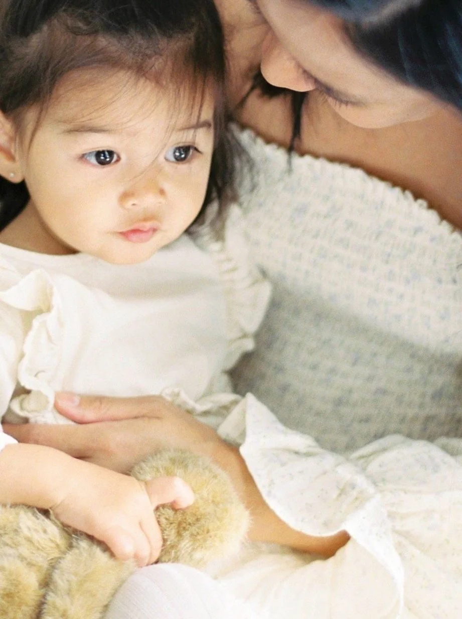 A young girl with dark hair and big eyes being gently held by an adult woman, possibly her mother, holding a small fluffy plush toy or animal, in a close, intimate moment.