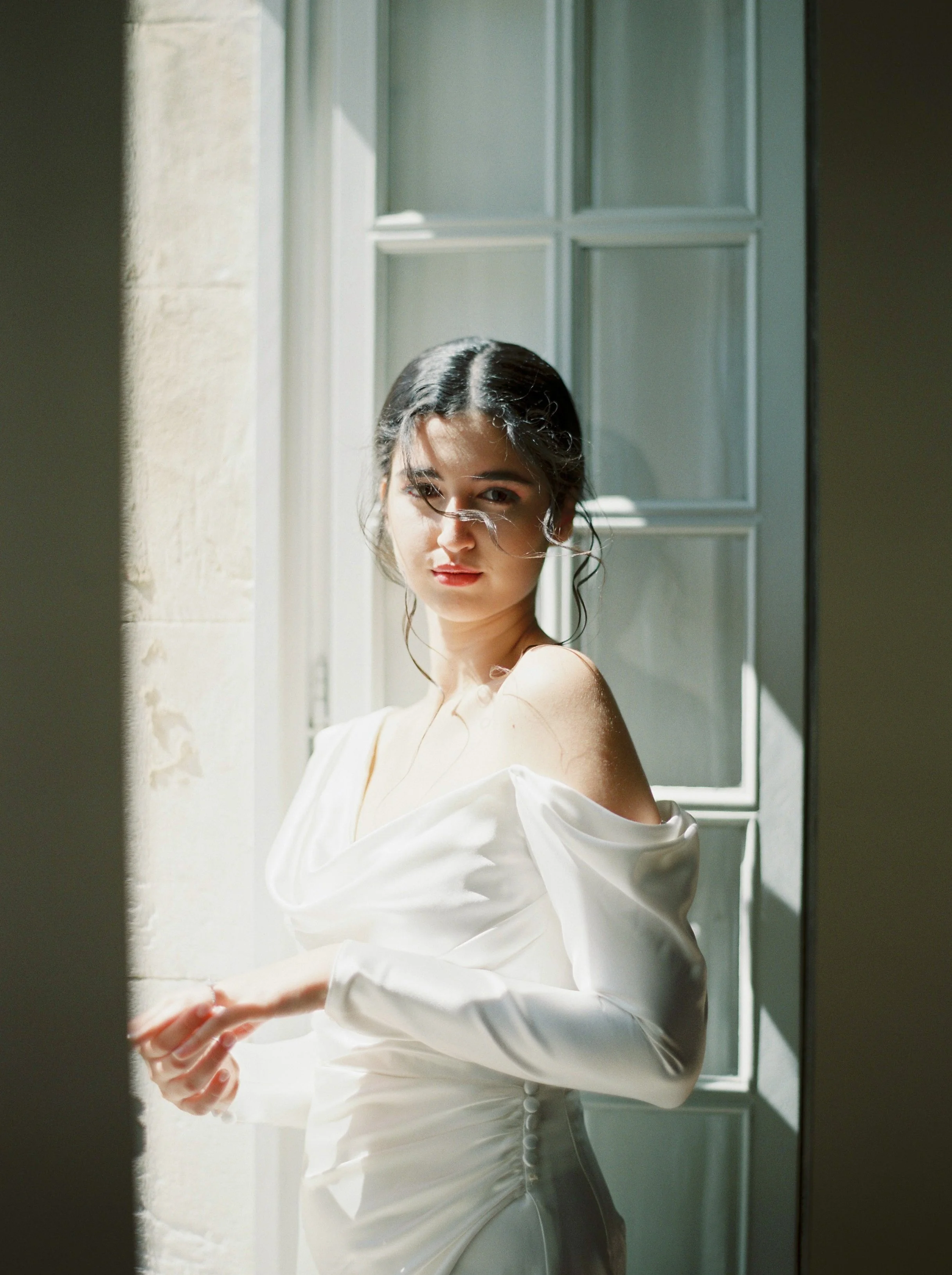 A woman in a white off-shoulder dress standing near a window with sunlight streaming in.