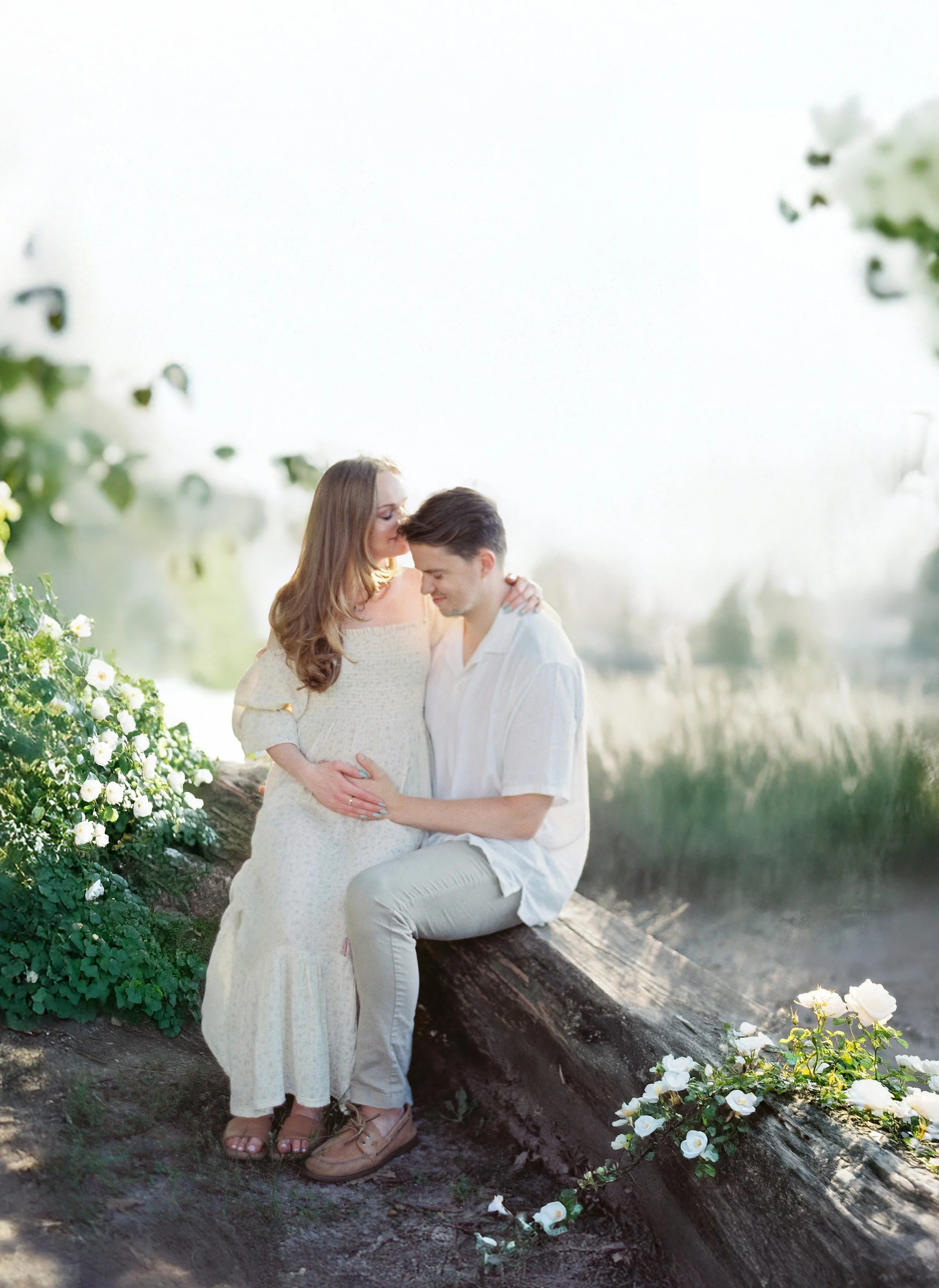 A pregnant woman and a man sitting on a log near a body of water, sharing an intimate moment in a nature setting with white flowers surrounding them.