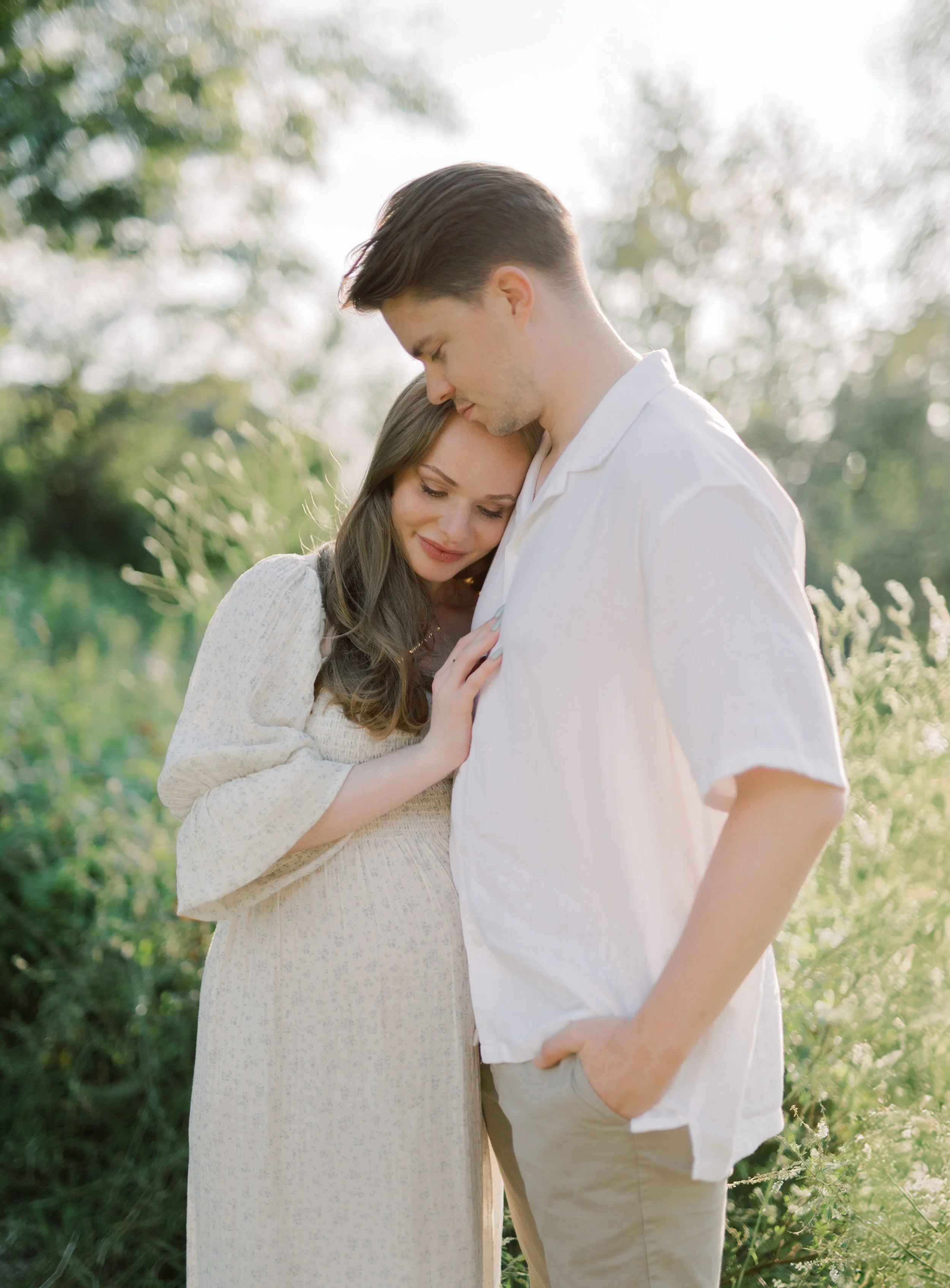 A pregnant woman and a man standing close together outside, with the woman's head resting on the man's chest and both smiling softly.