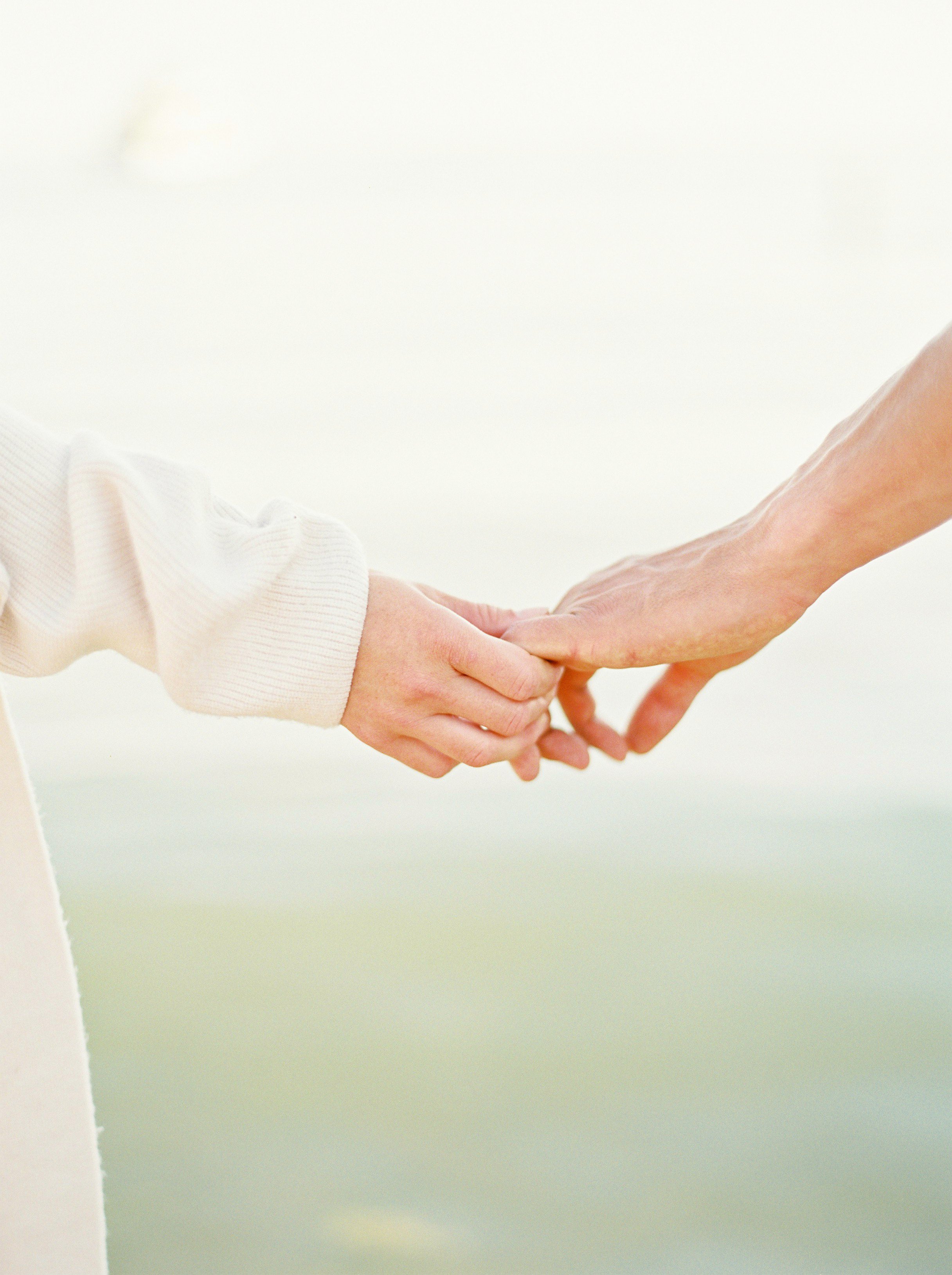 Two people holding hands, one with a white long sleeve shirt and the other with a bare arm, outdoors with a blurred bright background.