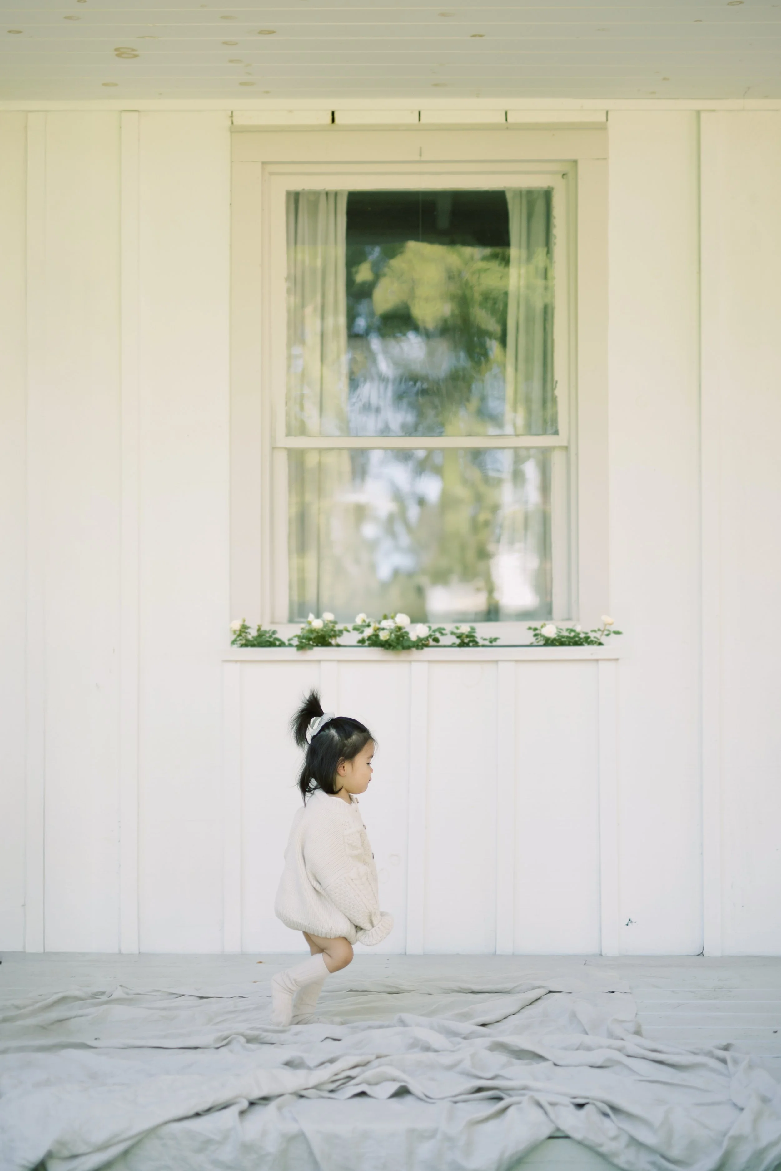 A young girl with dark hair in a ponytail, wearing a beige sweater and white socks, standing on a bed with white sheets in front of a white house wall and window with curtains and flowers.