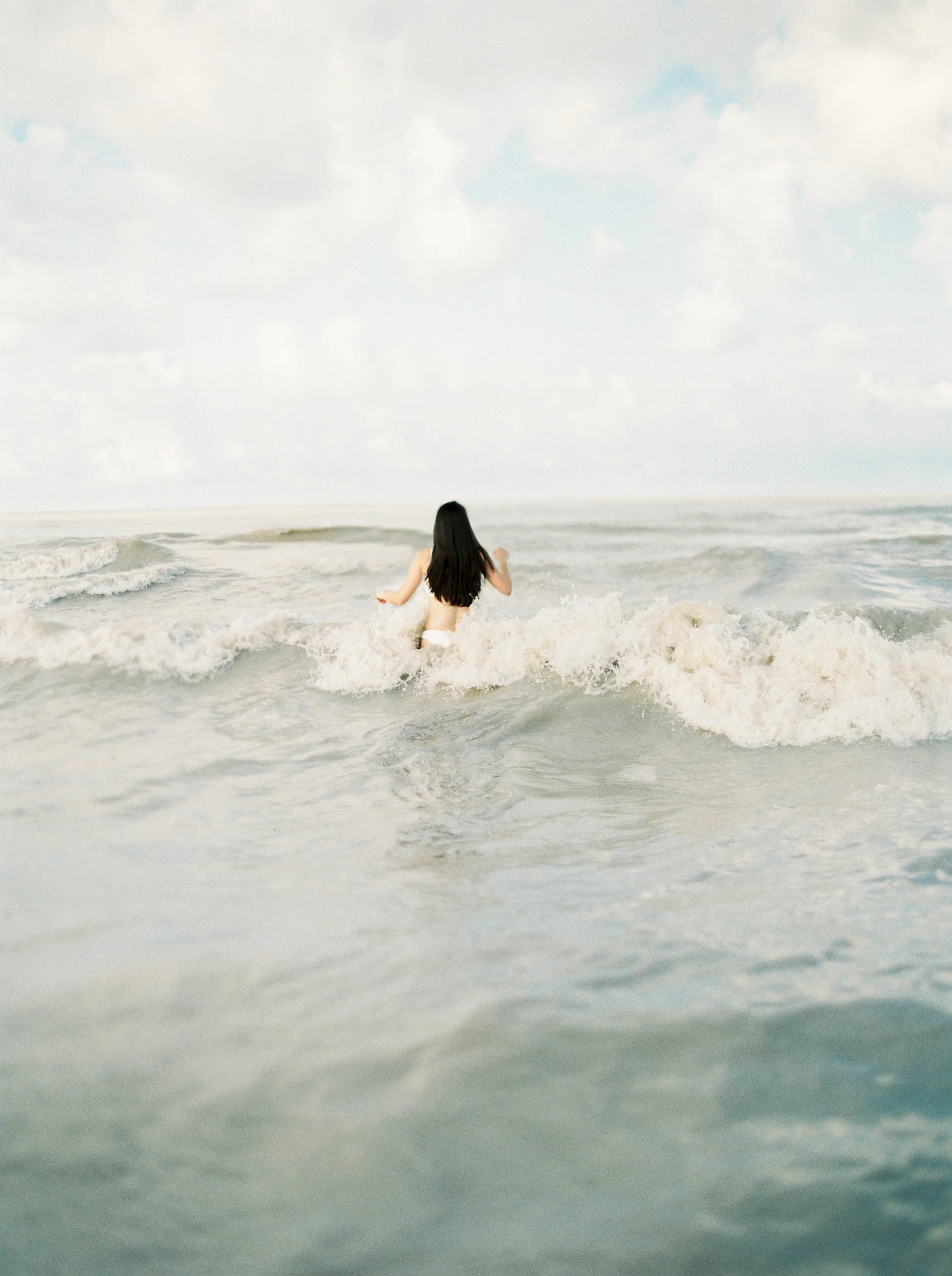 A woman with long dark hair, wearing a swimsuit, walking into the ocean waves on a cloudy day.