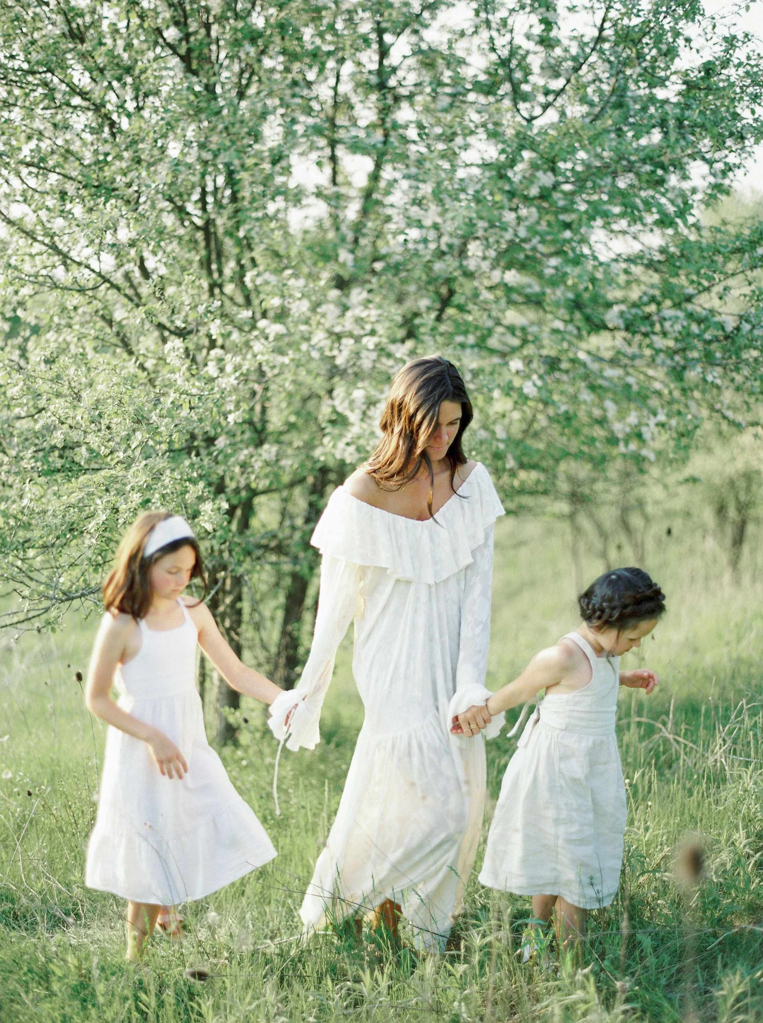 A woman and two young girls walking hand in hand through a grassy outdoor area with trees in the background. All three are wearing white dresses.