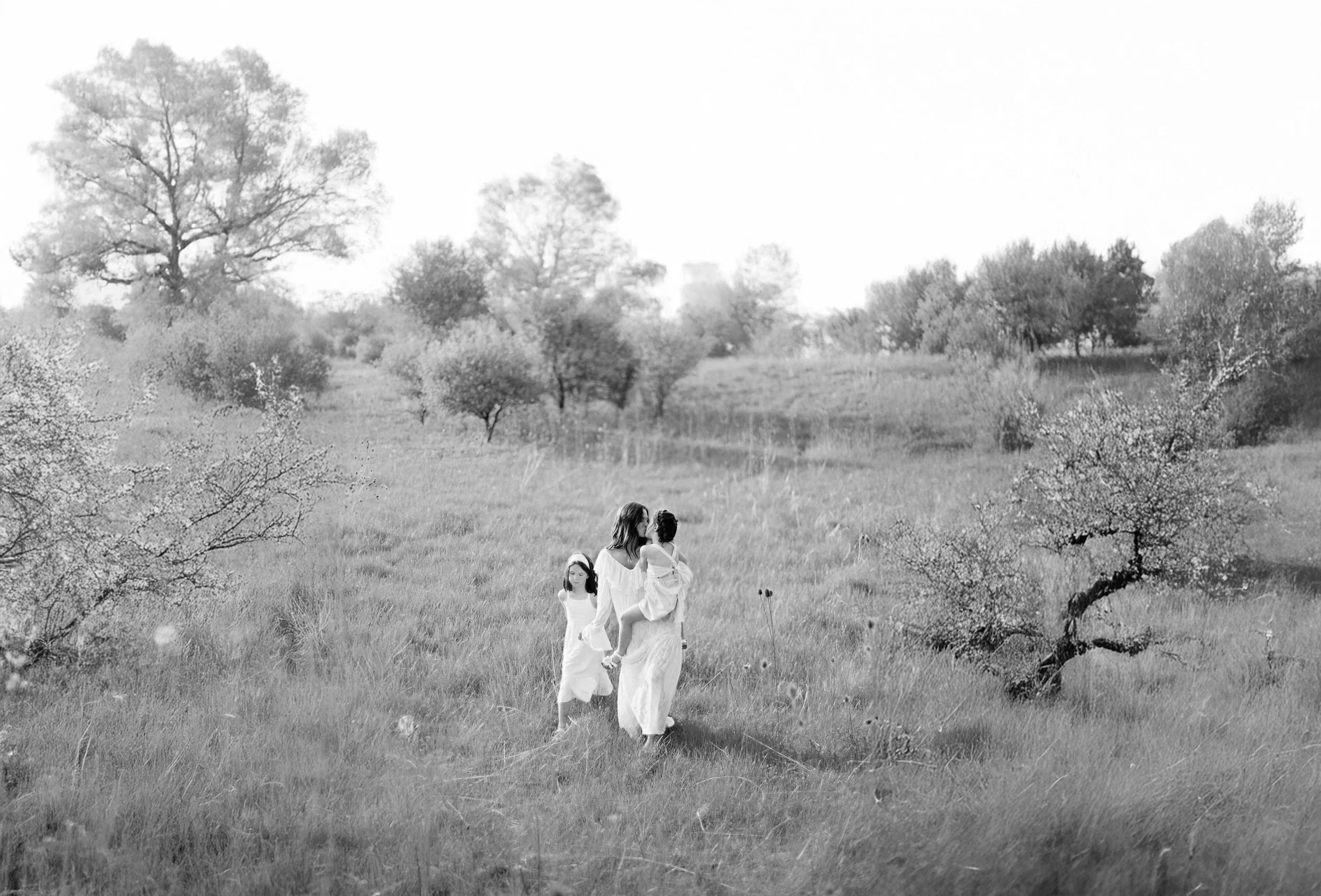A woman walking with two young girls through a grassy field with scattered trees, captured in black and white.