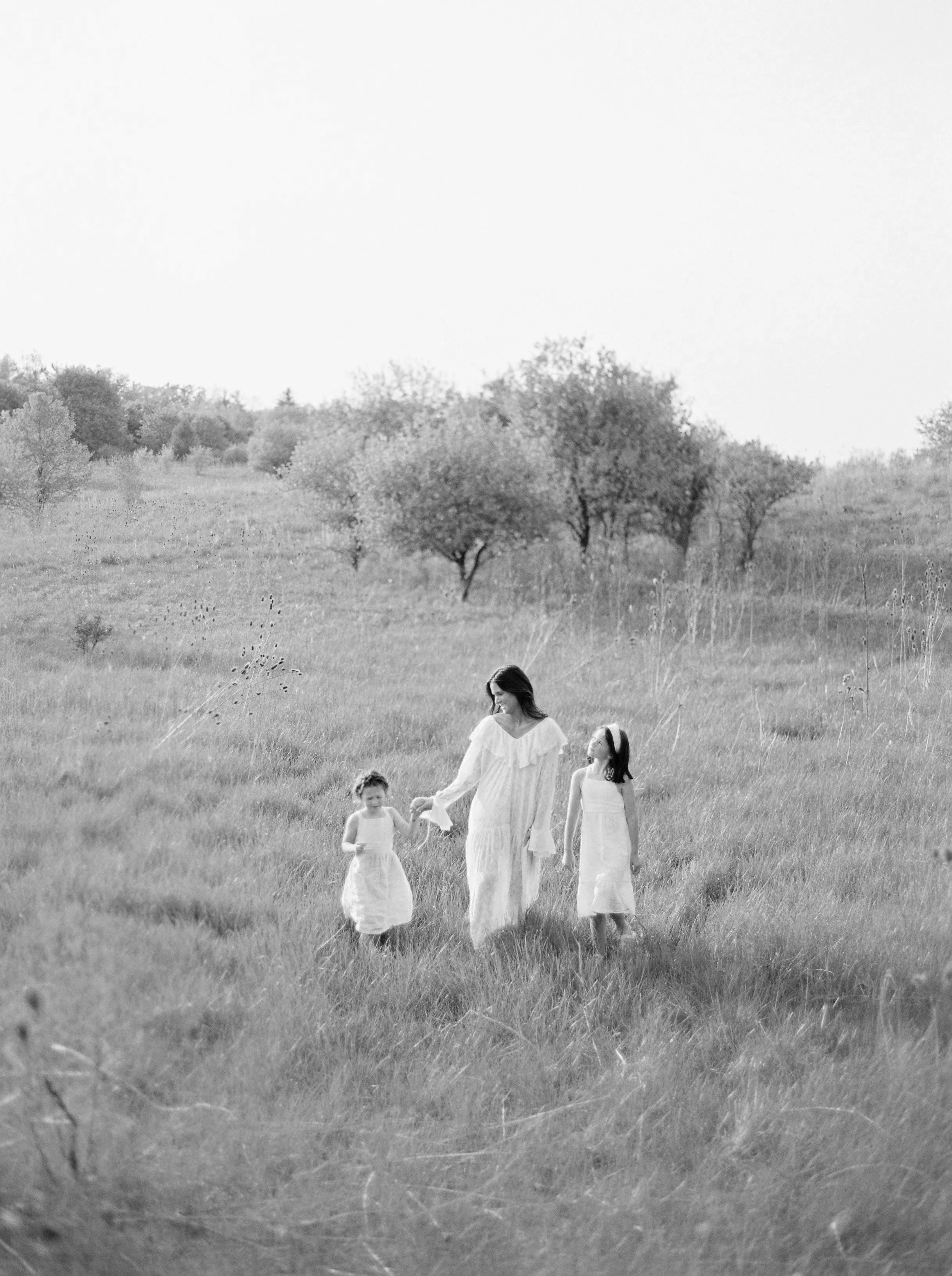A woman and two young girls walking through a grassy field with trees in the background, all dressed in white, black and white photo.