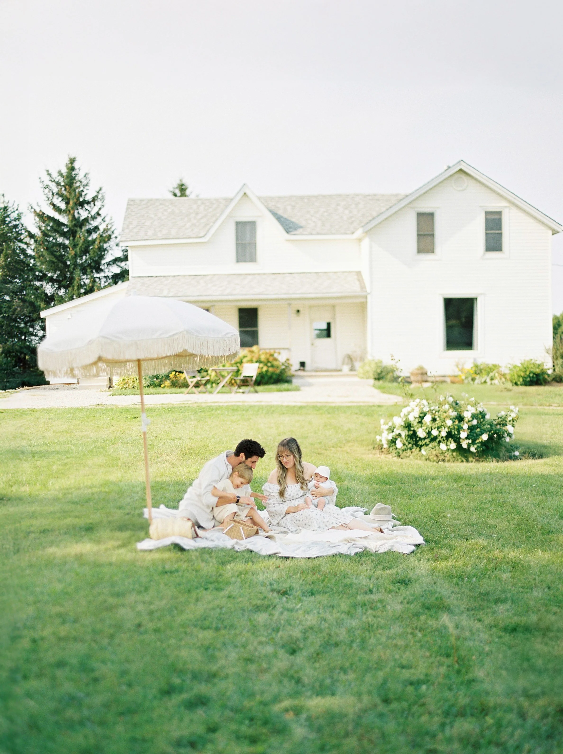 A family enjoying a picnic on a blanket in a lush backyard with a white house, umbrella, and garden in the background.